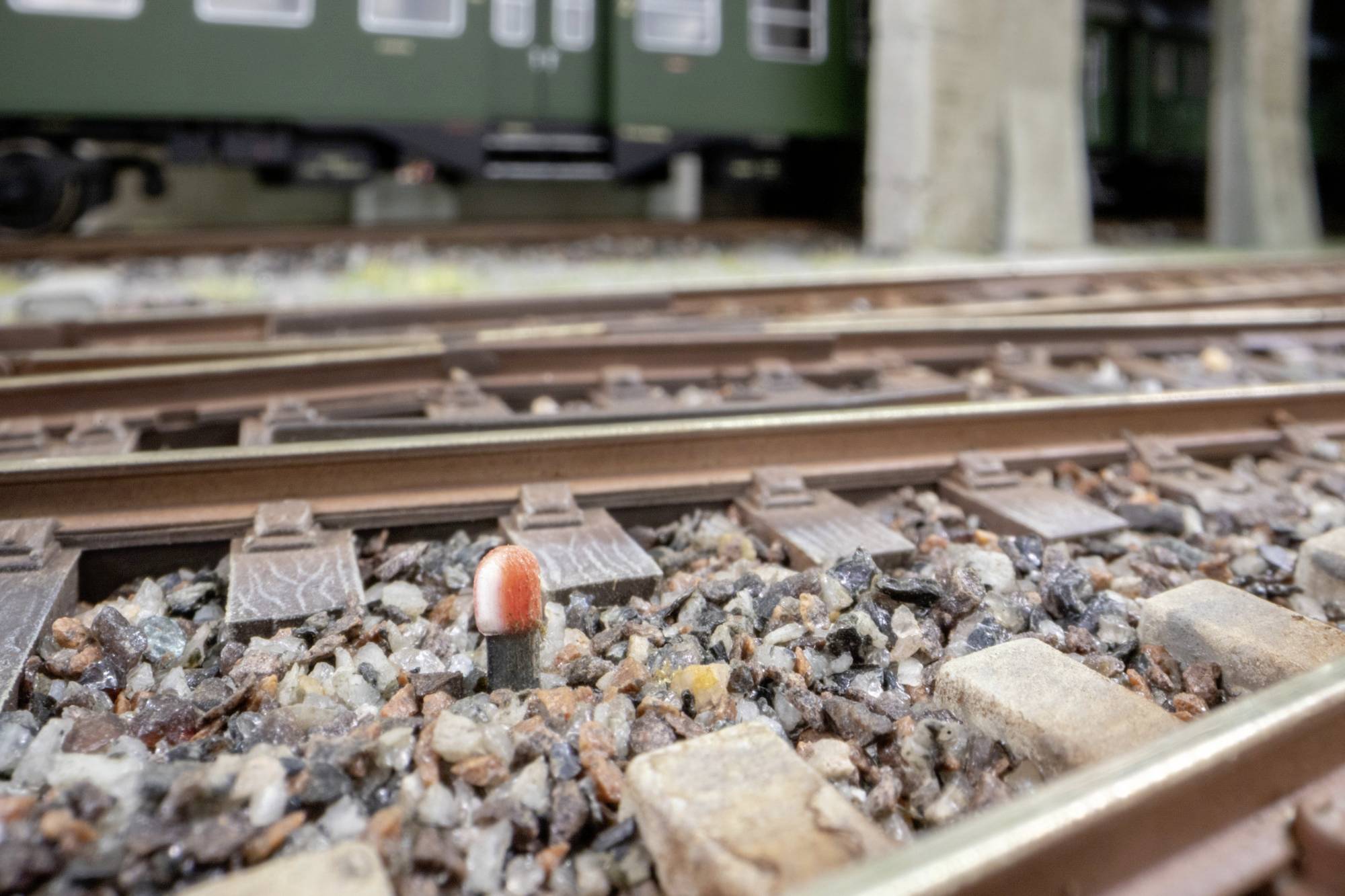 Primo piano dei binari del trenino in scala con ghiaia di ballast e un piccolo marcatore di binario rosso e bianco; carrozze del treno sfocate sullo sfondo.
