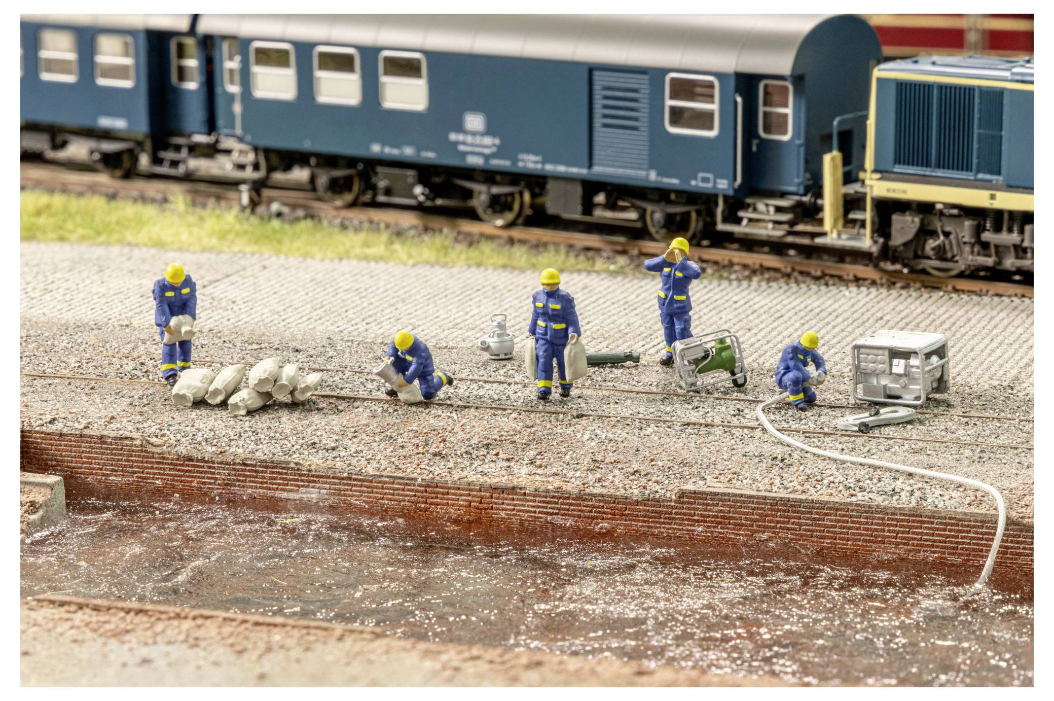 Scena di manutenzione della ferrovia in miniatura con minuscoli operai in caschi gialli e uniformi blu che riparano il binario accanto a un treno passeggeri blu.