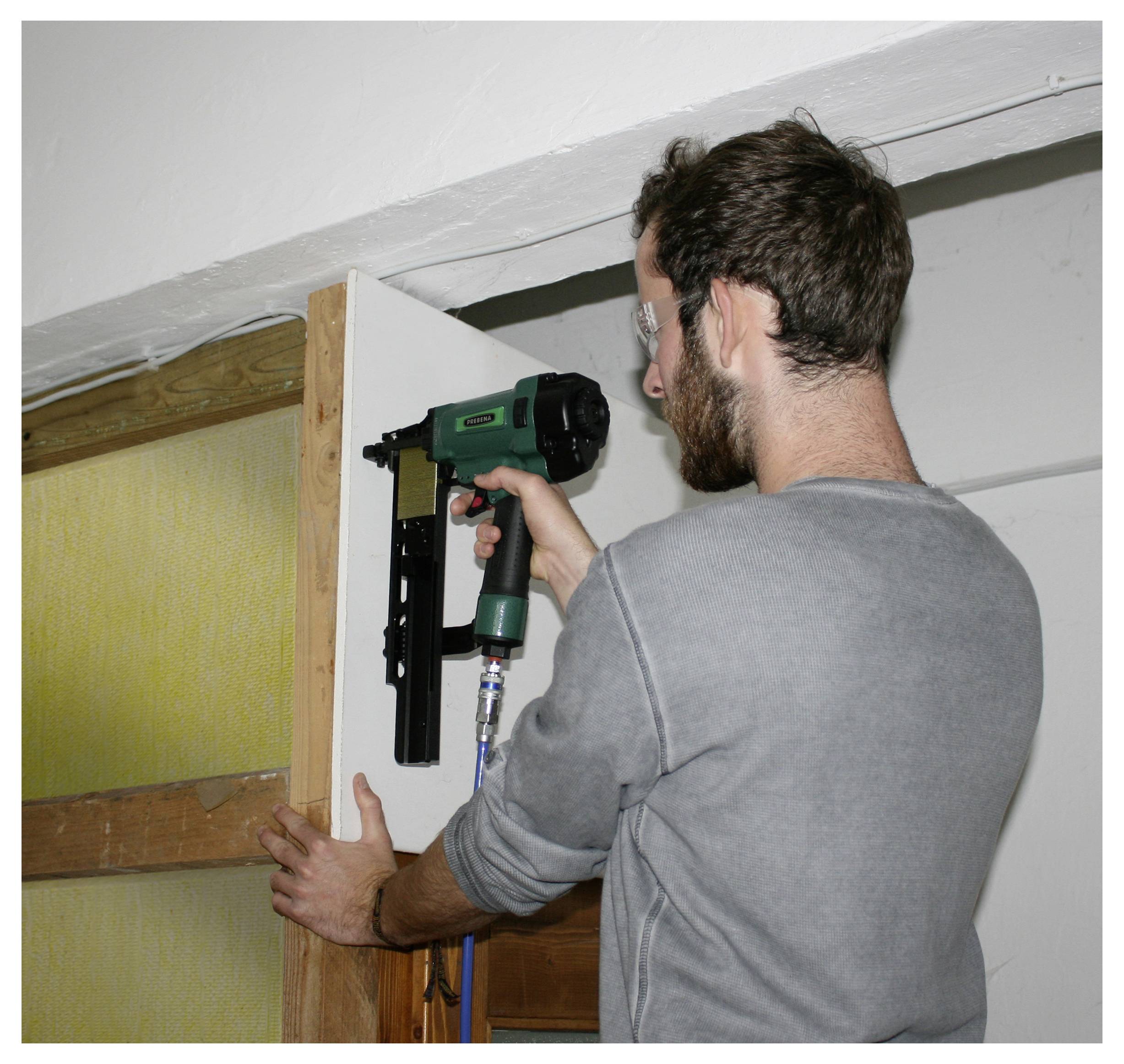 Uomo che utilizza una pistola chiodatrice pneumatica per installare un telaio di porta in legno all'interno di una stanza.
