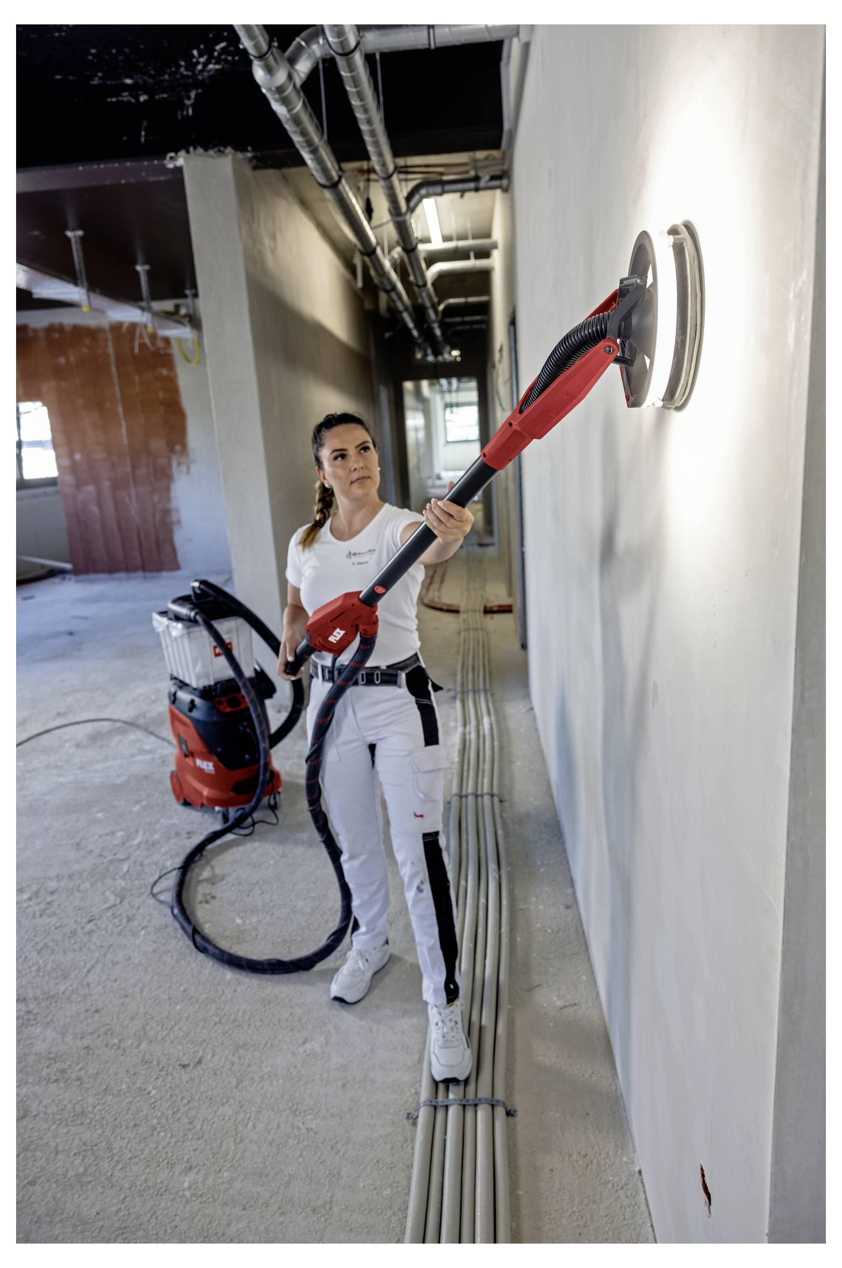 Un operaio leviga una parete con uno strumento a manico lungo in un corridoio di un edificio non finito, con un sistema di aspirazione sul pavimento dietro di lui.
