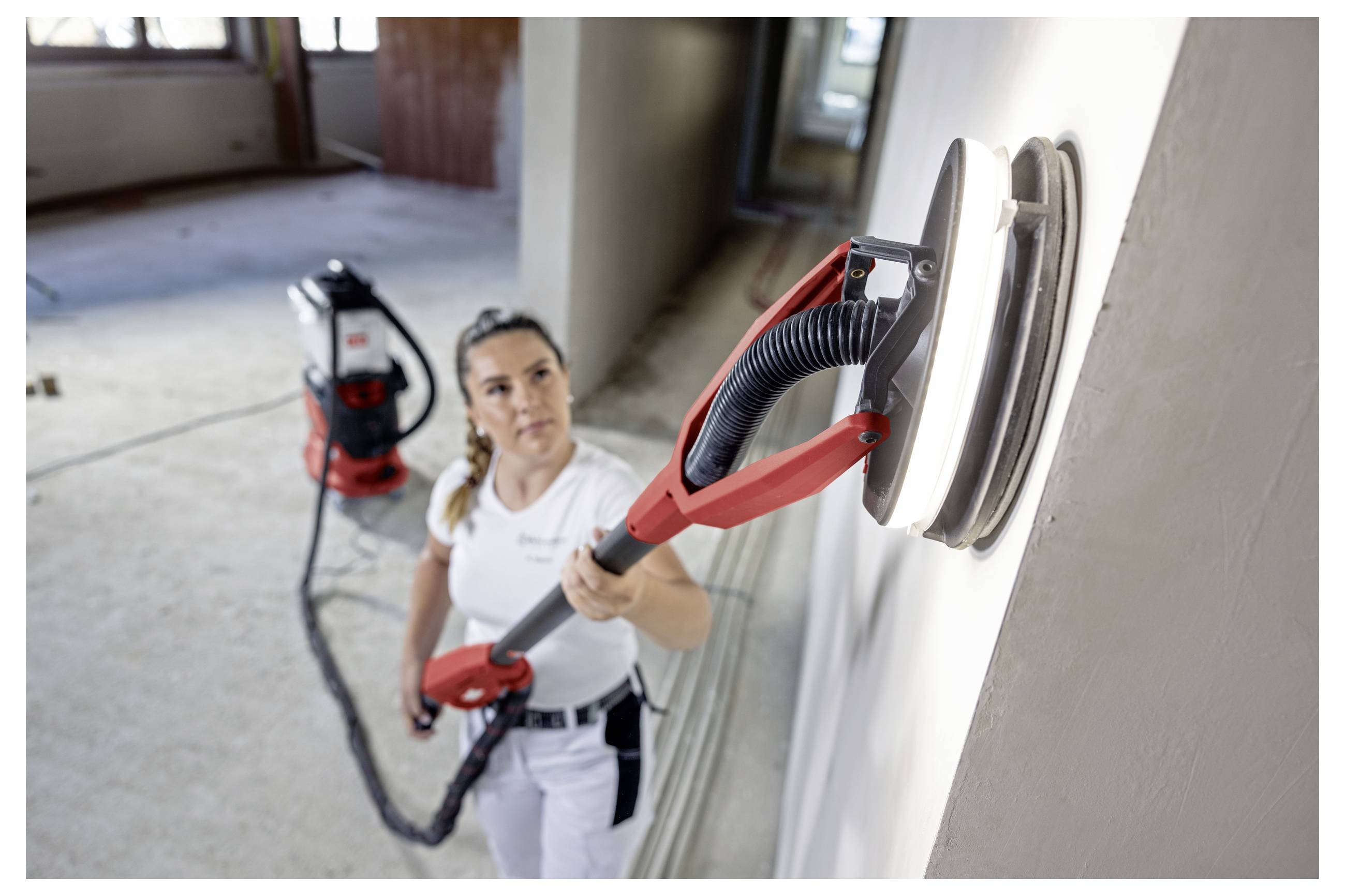 Un operaio utilizza una levigatrice a muro per cartongesso per levigare una parete interna, con un tubo di aspirazione collegato e un aspiratore per officina sullo sfondo.