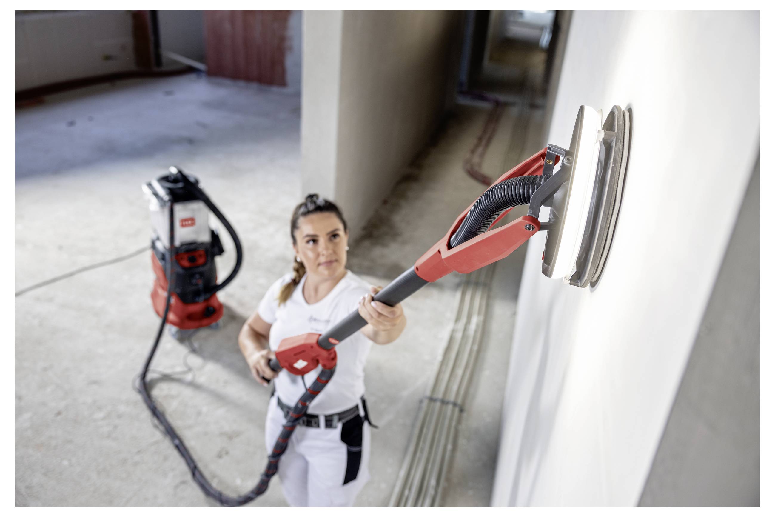 Donna che utilizza una levigatrice da muro rossa per levigare una parete interna durante la ristrutturazione della casa, con un estrattore di polvere a vuoto sullo sfondo.