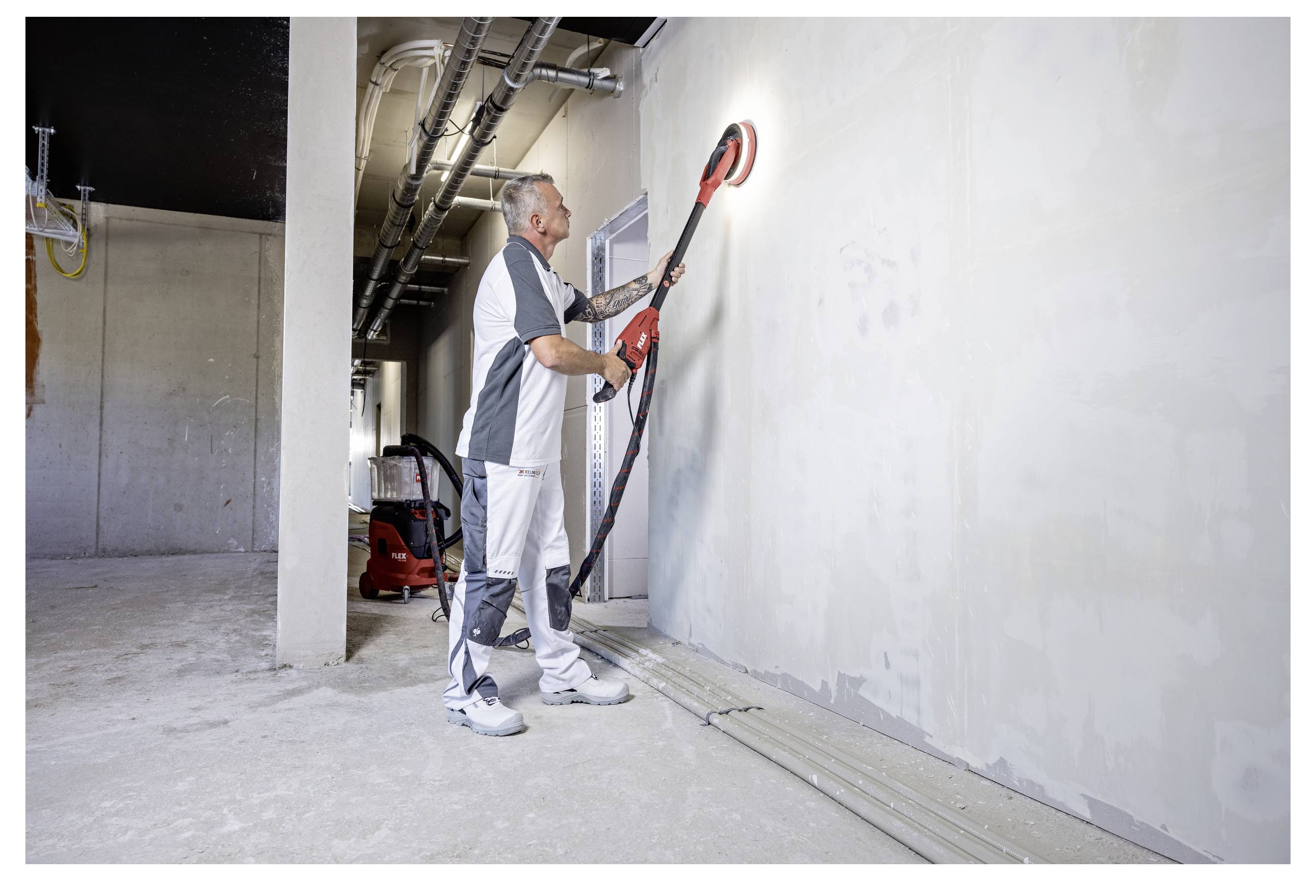 Un operaio utilizza una levigatrice elettrica a manico lungo su una parete all'interno di un edificio incompiuto, levigando la superficie vicino ai tubi del soffitto esposti.