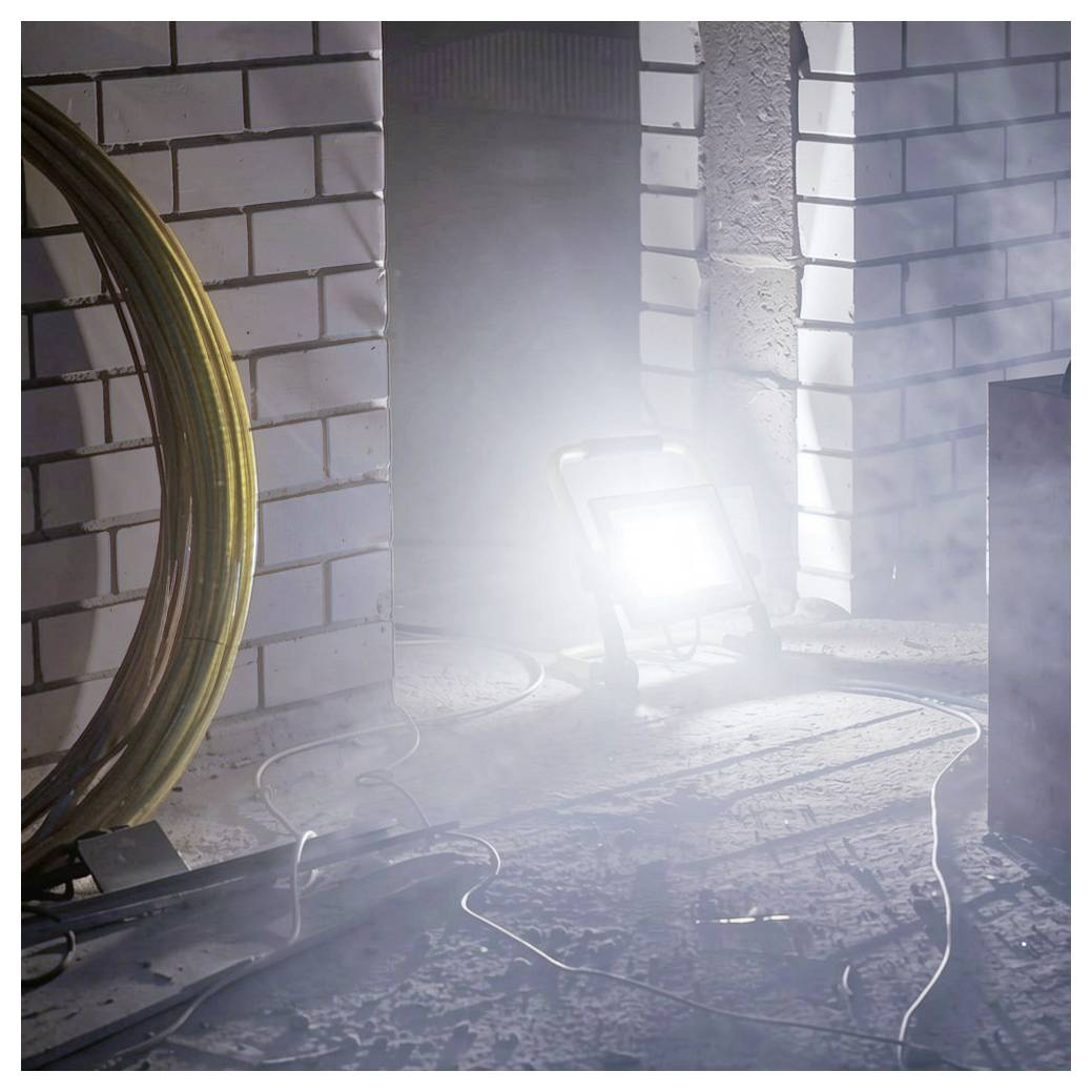 Lampada di lavoro luminosa sul pavimento di una stanza incompiuta e polverosa, con muri in laterizio, tubo arrotolato e cavi visibili.