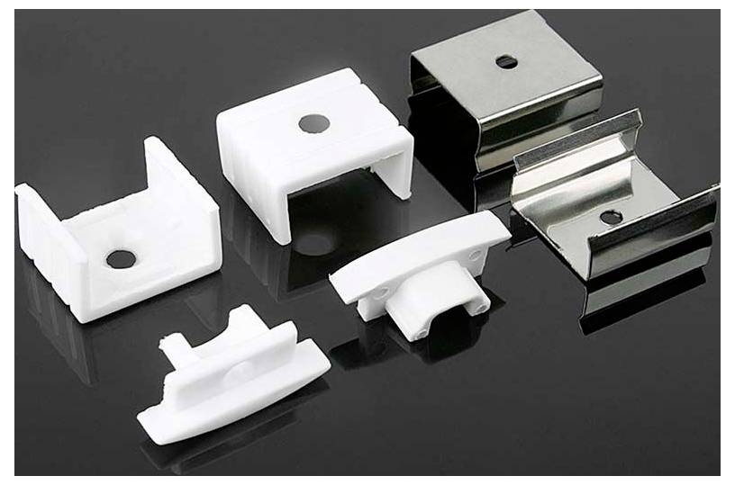 Diversi clip di montaggio o supporti in metallo e plastica disposti su una superficie scura, che mostrano diverse forme e finiture.
