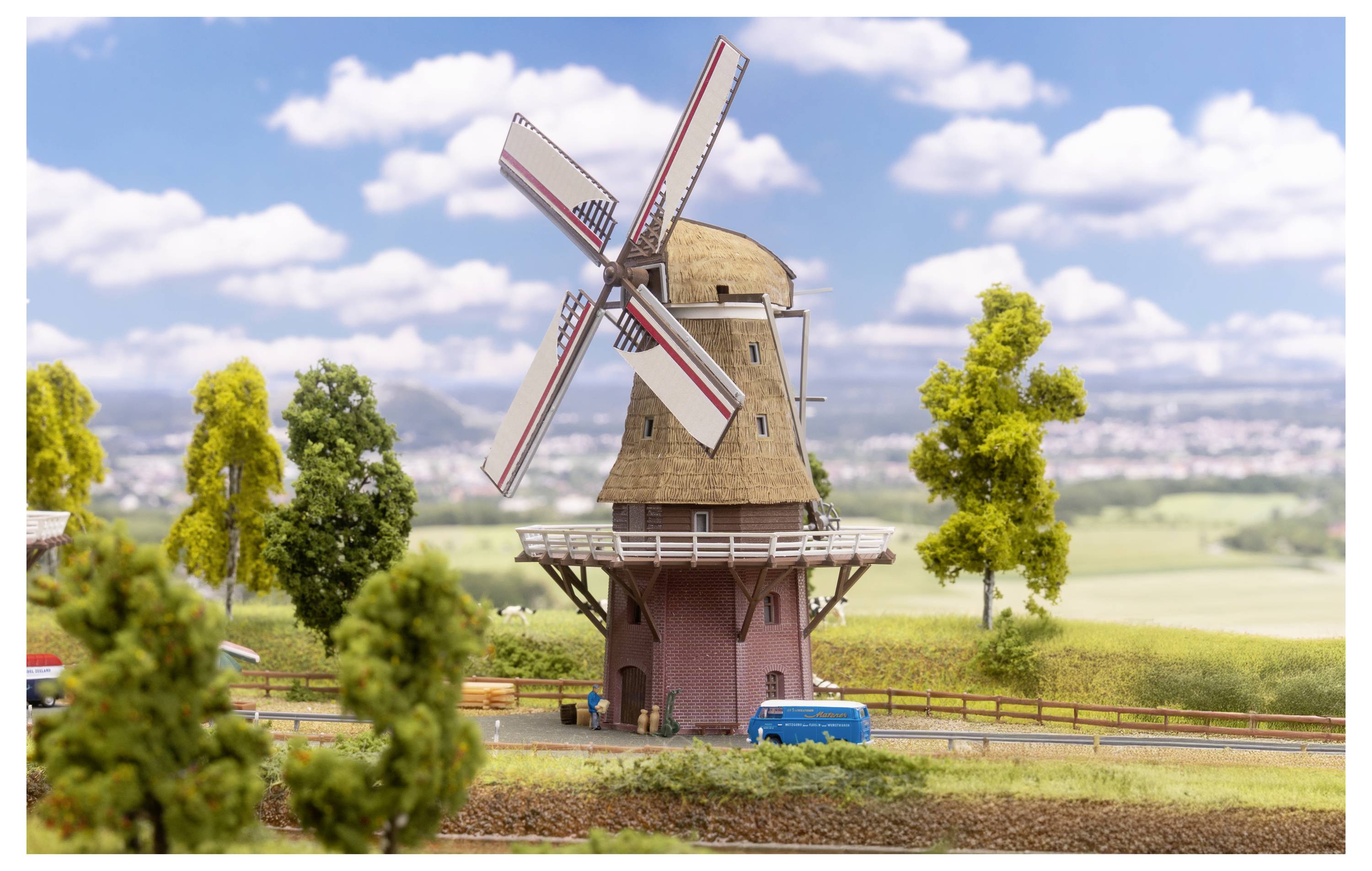 Mulino a vento in miniatura in un paesaggio rurale, con grandi pale, alberi, campi e un cielo blu con nuvole.