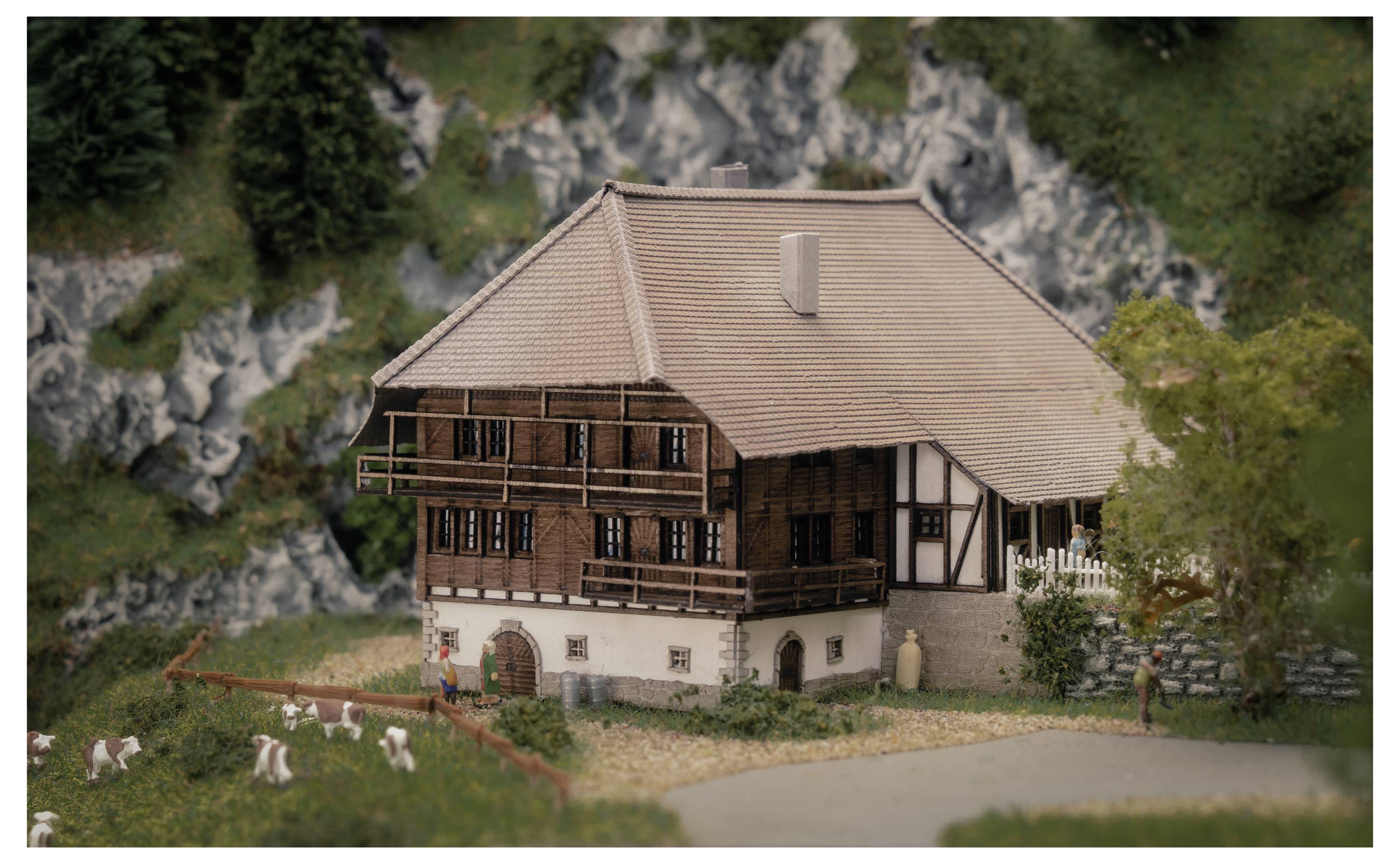 Un modello di una rustica casa colonica a due piani con tetto in tegole, posizionata accanto a un recinto con un giardino erboso contenente piccoli animali al pascolo e sullo sfondo un paesaggio di colline rocciose.