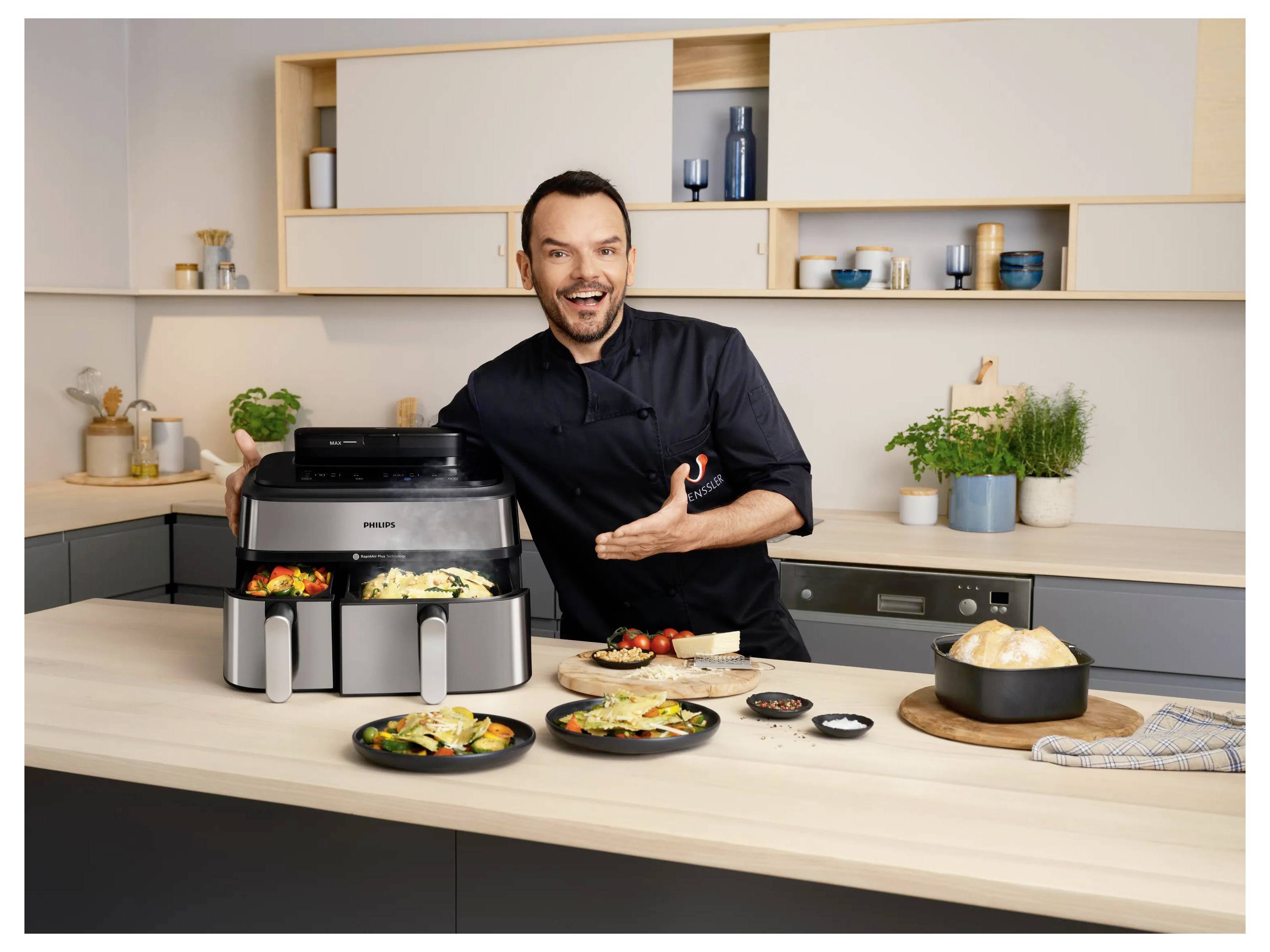 Un uomo sorridente presenta una friggitrice ad aria Philips con cibo cotto in una cucina moderna, circondato da piatti pronti e una ciotola di riso.