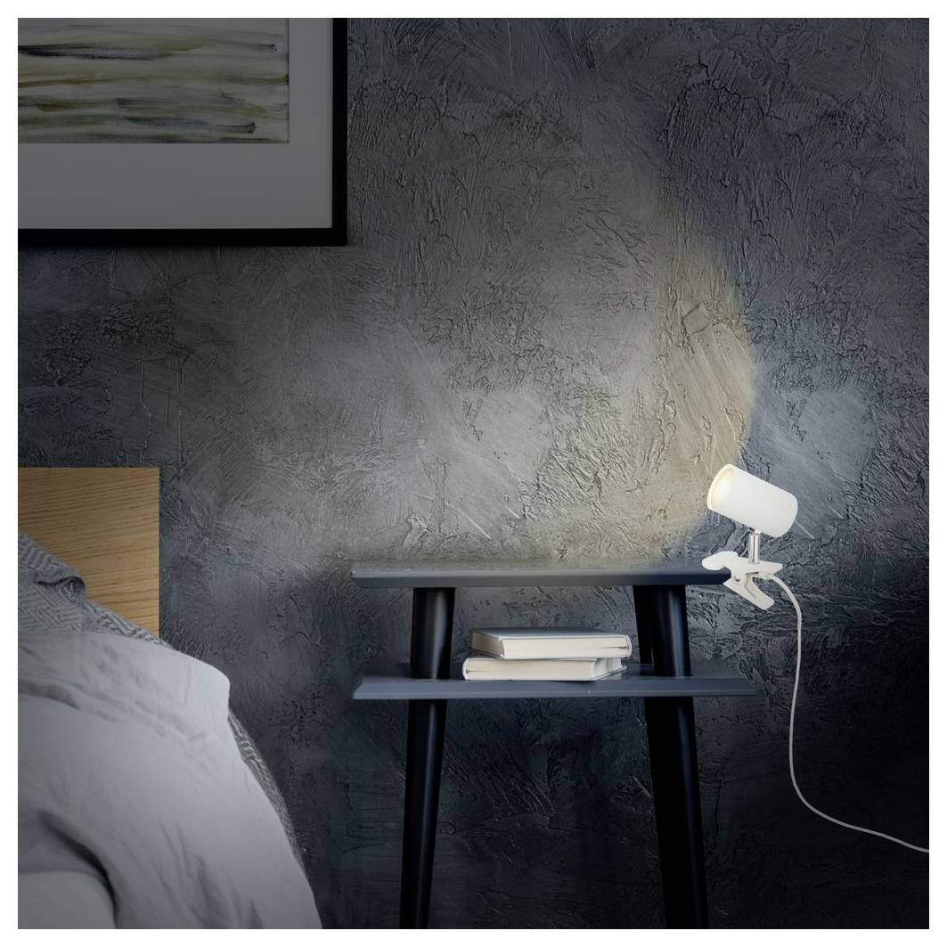 Scena di una camera da letto in penombra con un comodino, libri impilati e una lampada regolabile accesa contro un muro grigio strutturato accanto a un letto.