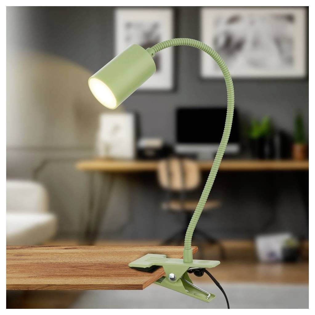 Lampada da scrivania verde a clip con collo flessibile, agganciata a un tavolo in legno e accesa su uno sfondo sfocato di un ufficio domestico.
