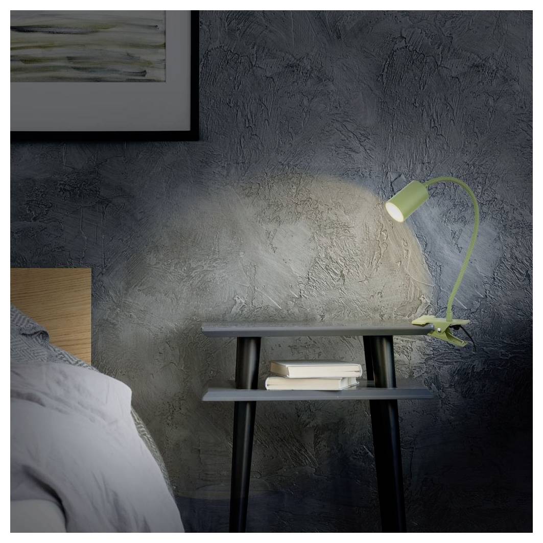 Un comodino con una lampada verde e libri accanto a un letto contro un muro dalla trama scura.