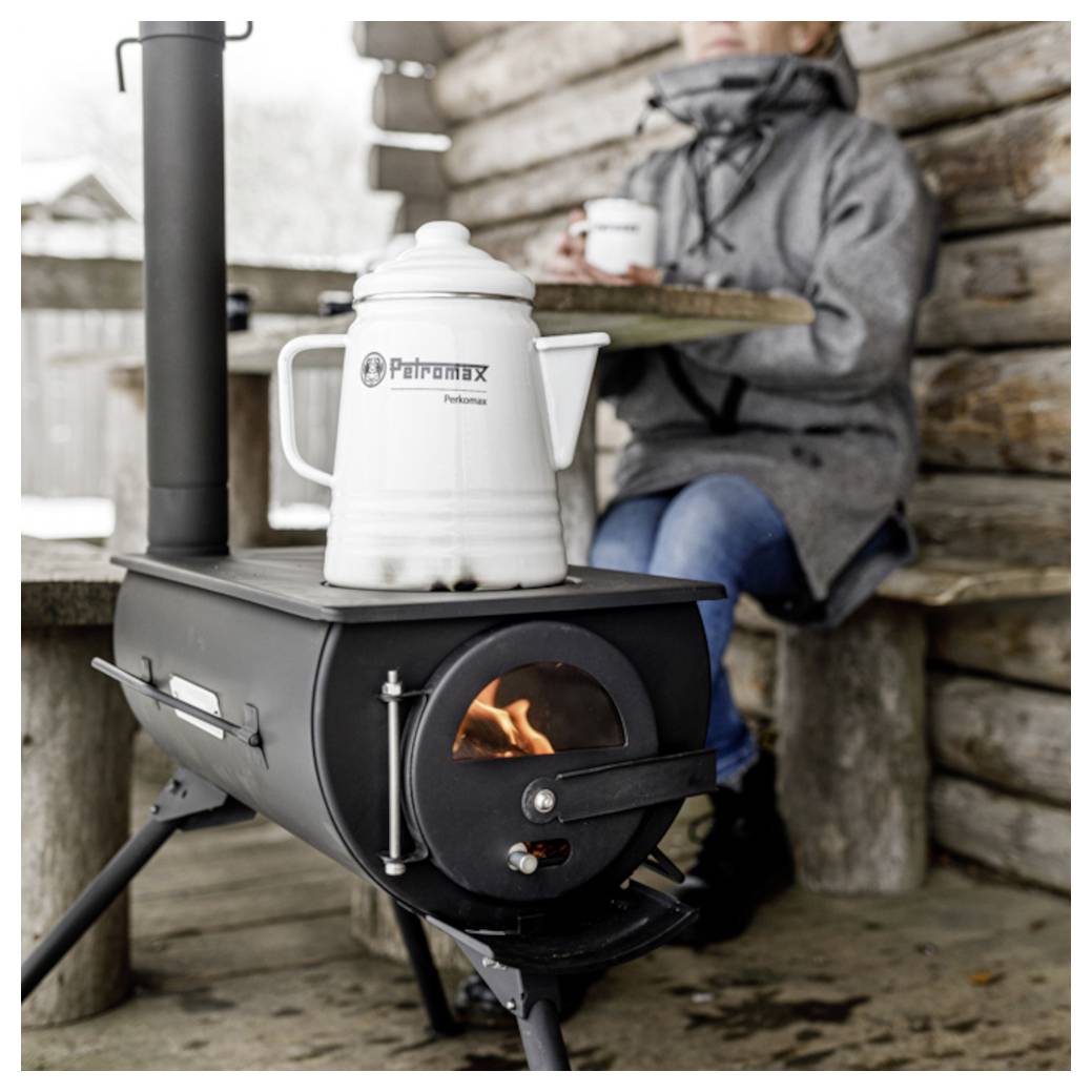 Una stufa a legna riscalda un bollitore Petromax bianco mentre una persona in un cappotto invernale siede nelle vicinanze tenendo una tazza su un portico rustico.