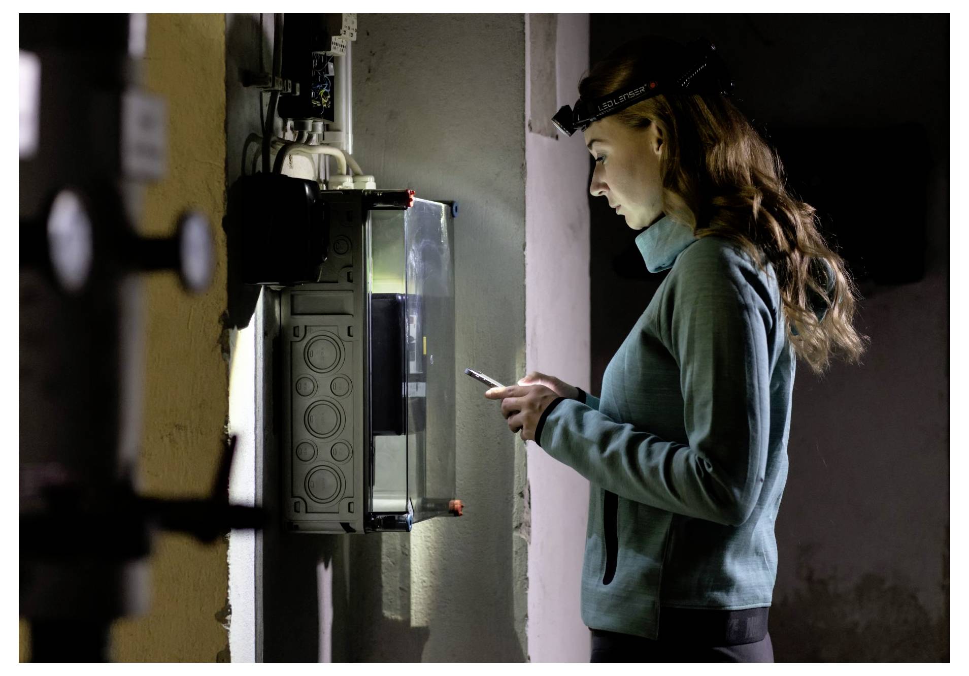 Una donna che indossa una lampada frontale controlla il suo telefono accanto a un pannello elettrico aperto in una zona di servizio poco illuminata.