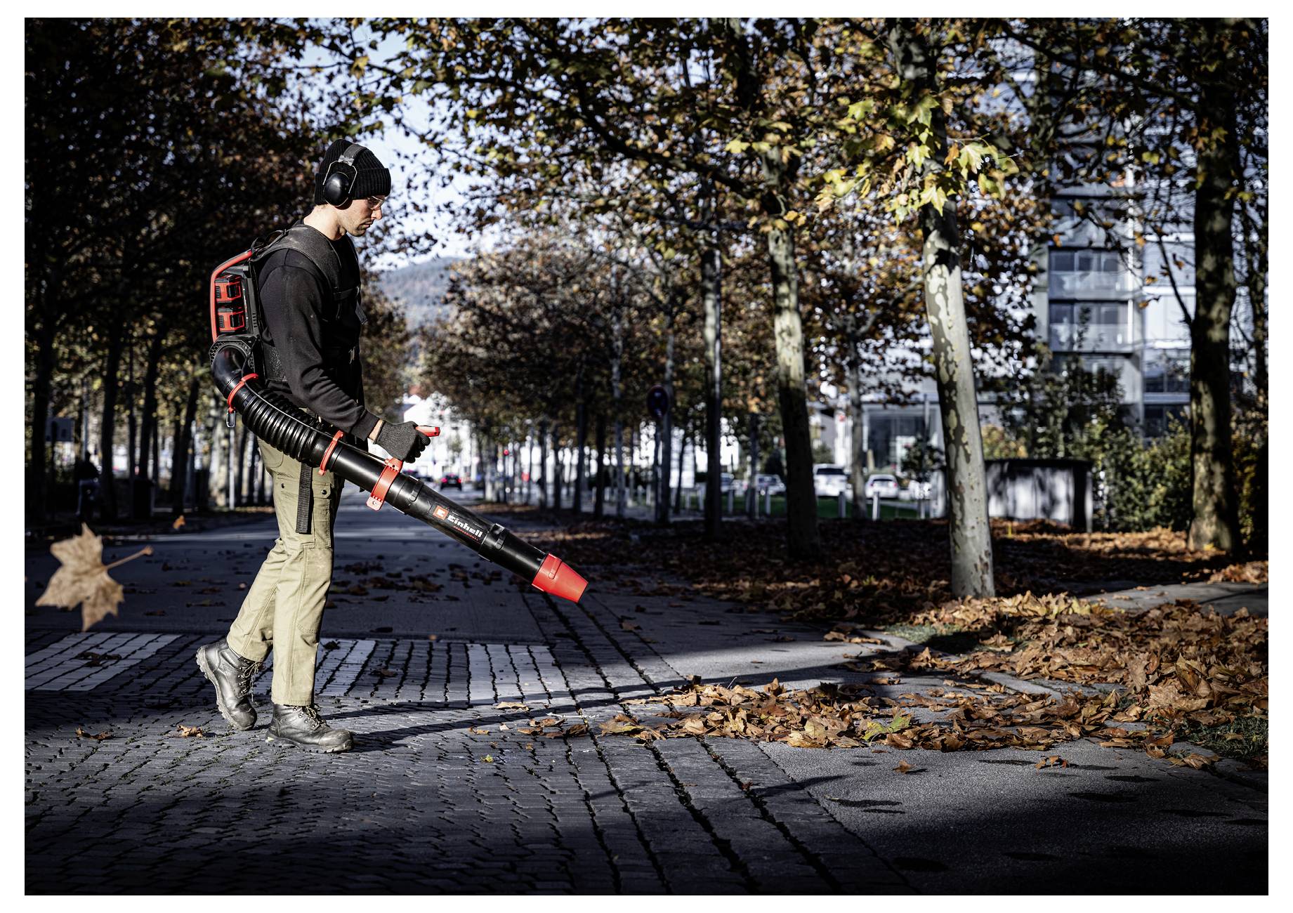 Un operaio che utilizza un soffiatore di foglie a zaino per ripulire le foglie cadute da un marciapiede alberato in autunno.