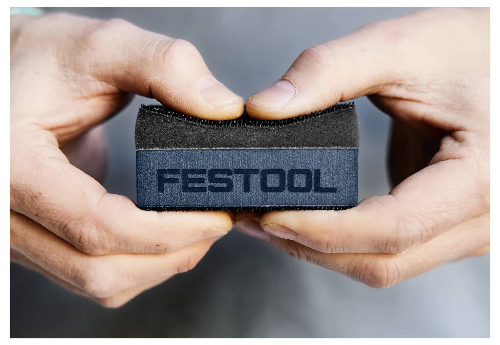 Due mani tengono un piccolo accessorio o strumento marchiato 'FESTOOL' di colore nero e blu contro uno sfondo sfocato.