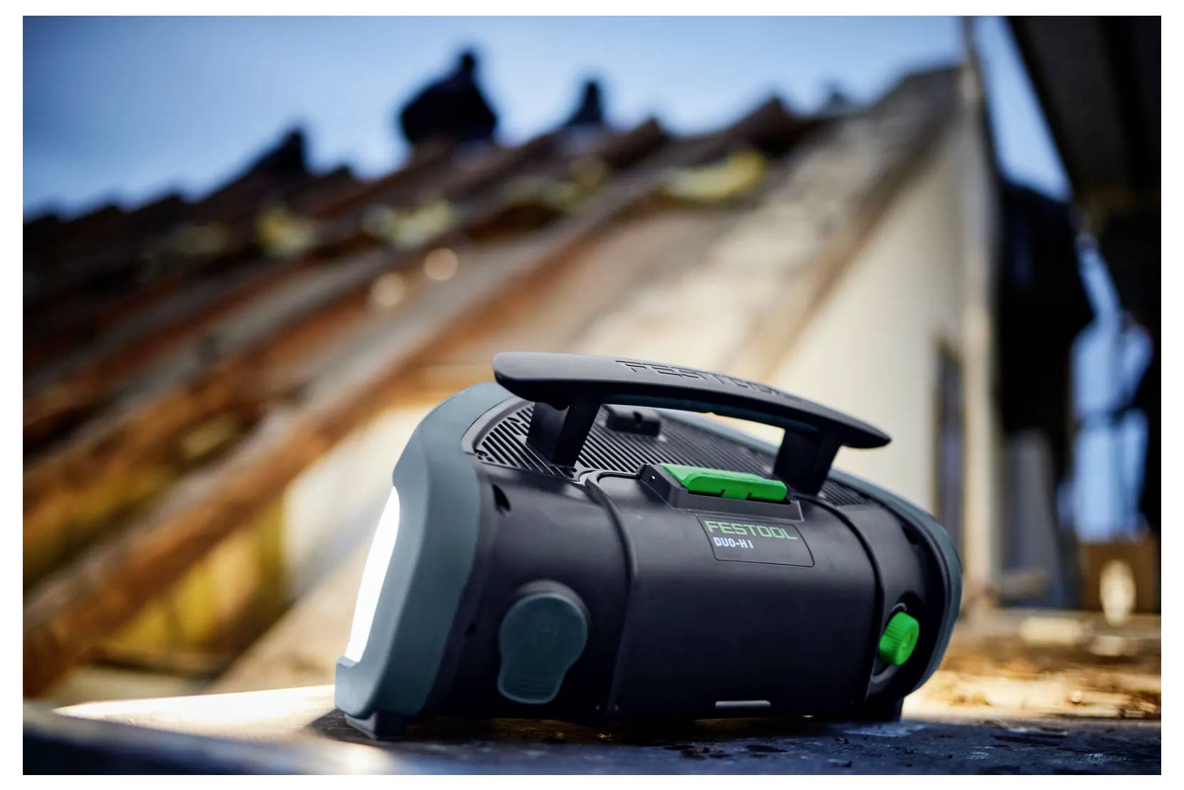 Radio portatile nera Festool senza batteria/utensile su una superficie di lavoro, con un tetto sfocato e un operaio sullo sfondo.