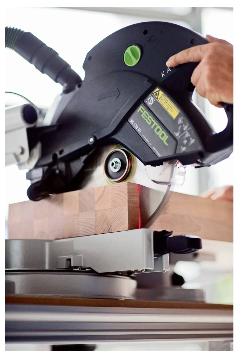 Primo piano di una sega a troncatura Festool Kapex che taglia un blocco di legno impilato, con una mano che preme il pulsante di controllo della sega.