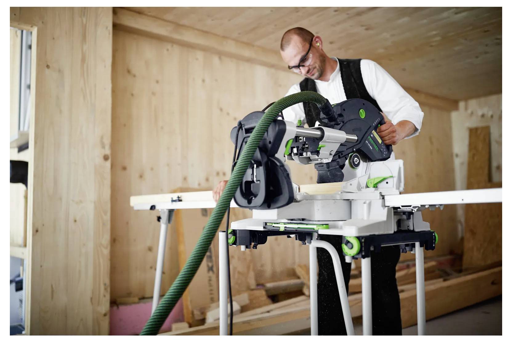 Uomo che usa una sega a spalla per tagliare il legno all'interno di una stanza in legno non rifinita, con un tubo di raccolta della polvere collegato.