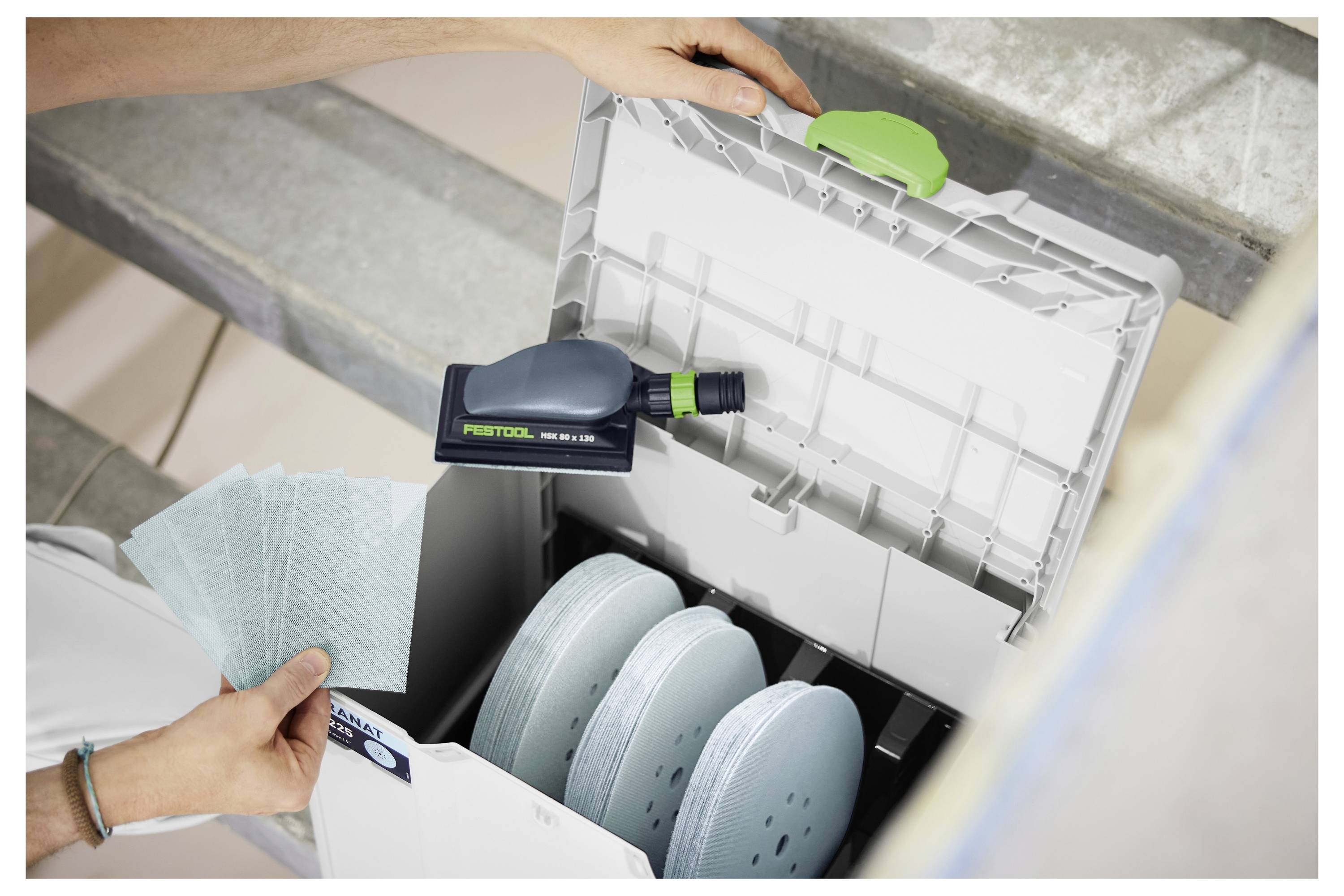 Mani che inseriscono dischi abrasivi in una scatola di stoccaggio Festool Systainer con un utensile elettrico allegato all'interno.