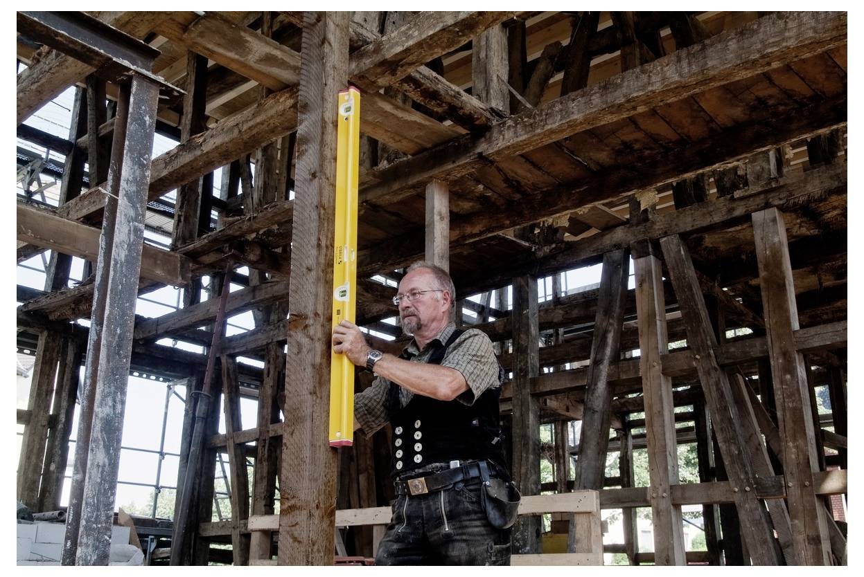 Un uomo in abiti da lavoro utilizza una livella a bolla per controllare una trave di legno all'interno di una struttura in legno antico parzialmente costruita.