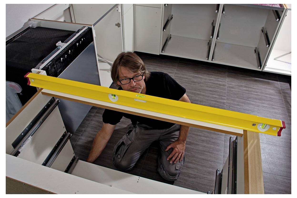 Un uomo inginocchiato all'interno di un'installazione di un mobile cucina, controlla l'allineamento con una livella gialla attraverso il telaio del piano di lavoro.