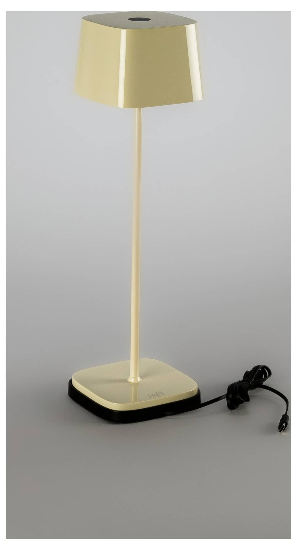 Lampada da tavolo in oro con paralume conico, stelo sottile, base quadrata e cavo di alimentazione nero su sfondo grigio.