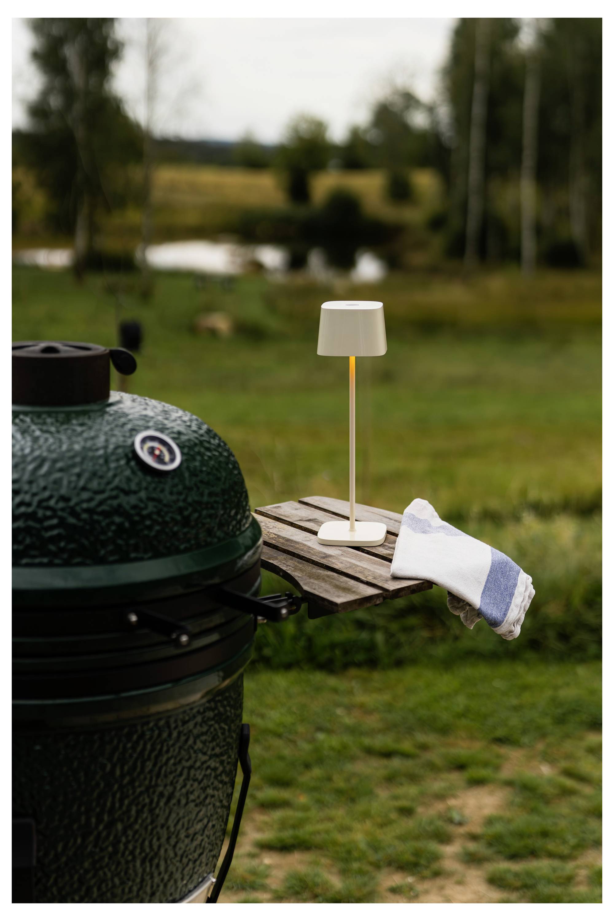 Una griglia in ceramica verde accanto a un piccolo tavolo laterale in legno che sostiene una lampada bianca e un asciugamano piegato, posizionata su un patio erboso con uno stagno sfocato e alberi sullo sfondo.