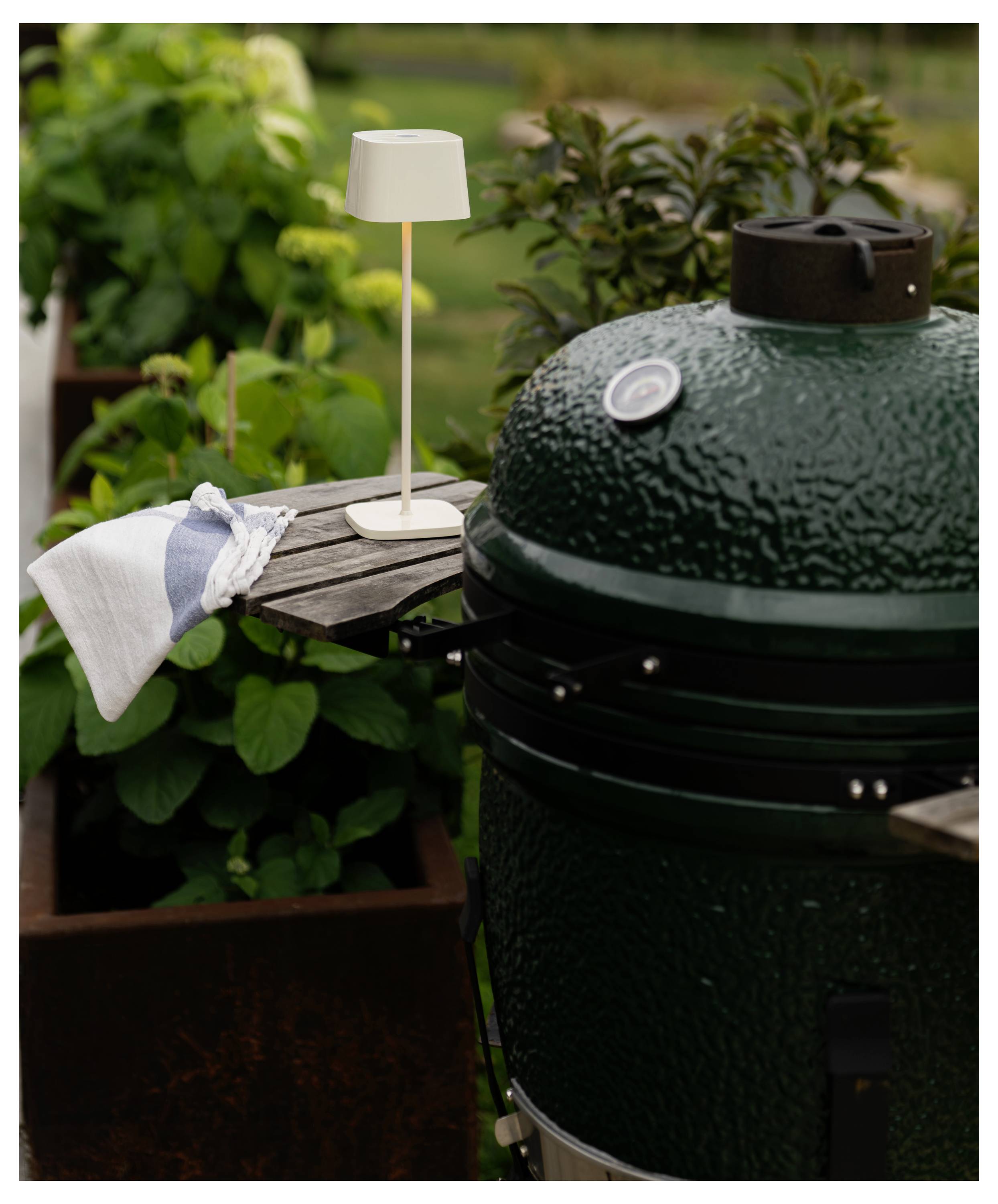 Griglia ceramica verde all'aperto accanto a un piccolo tavolino laterale con lampada, asciugamano e piante in vaso su un patio.