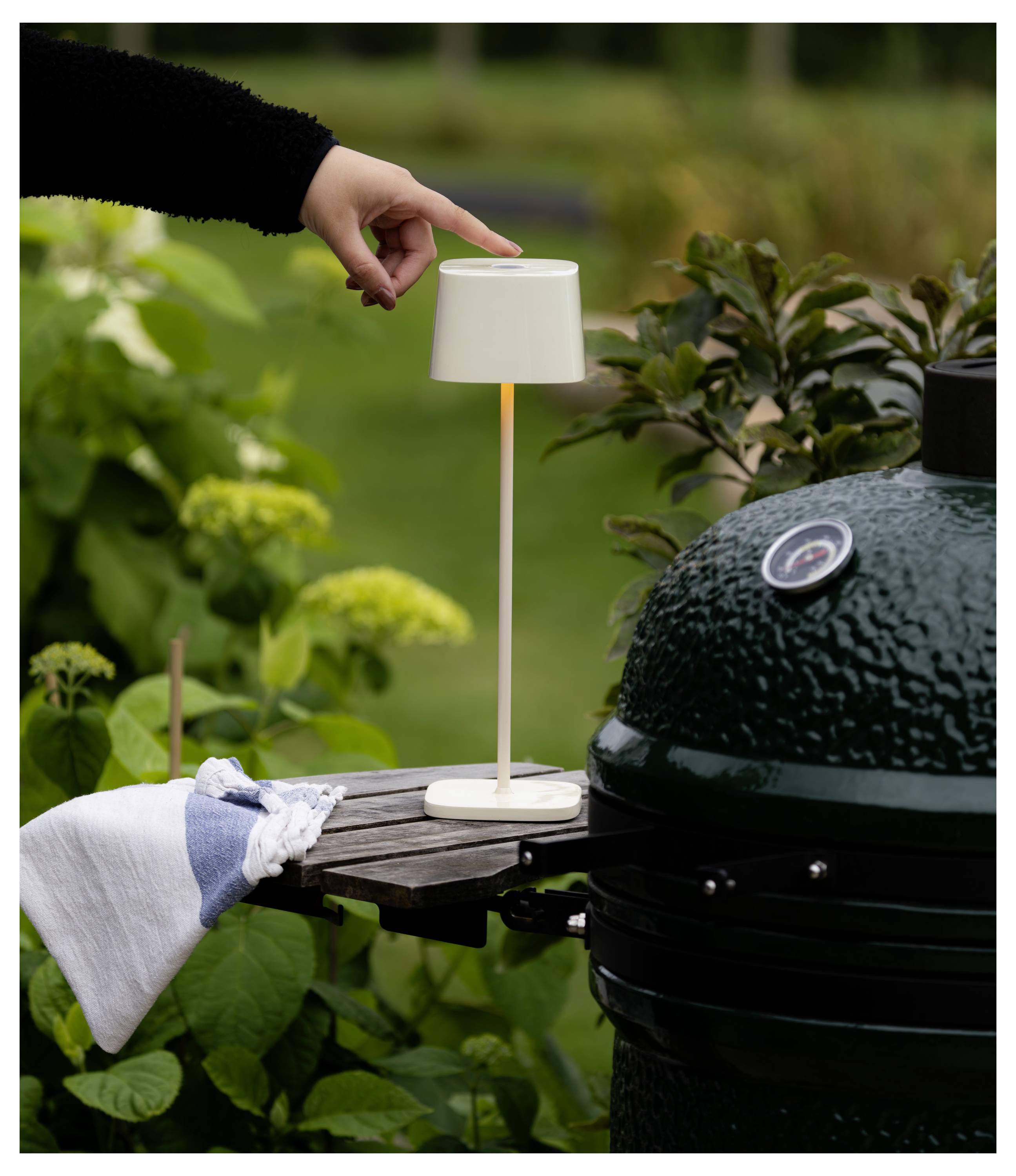 Una mano tocca leggermente una piccola lampada da tavolo wireless su uno scaffale laterale di una griglia in un'ambientazione di patio da giardino.