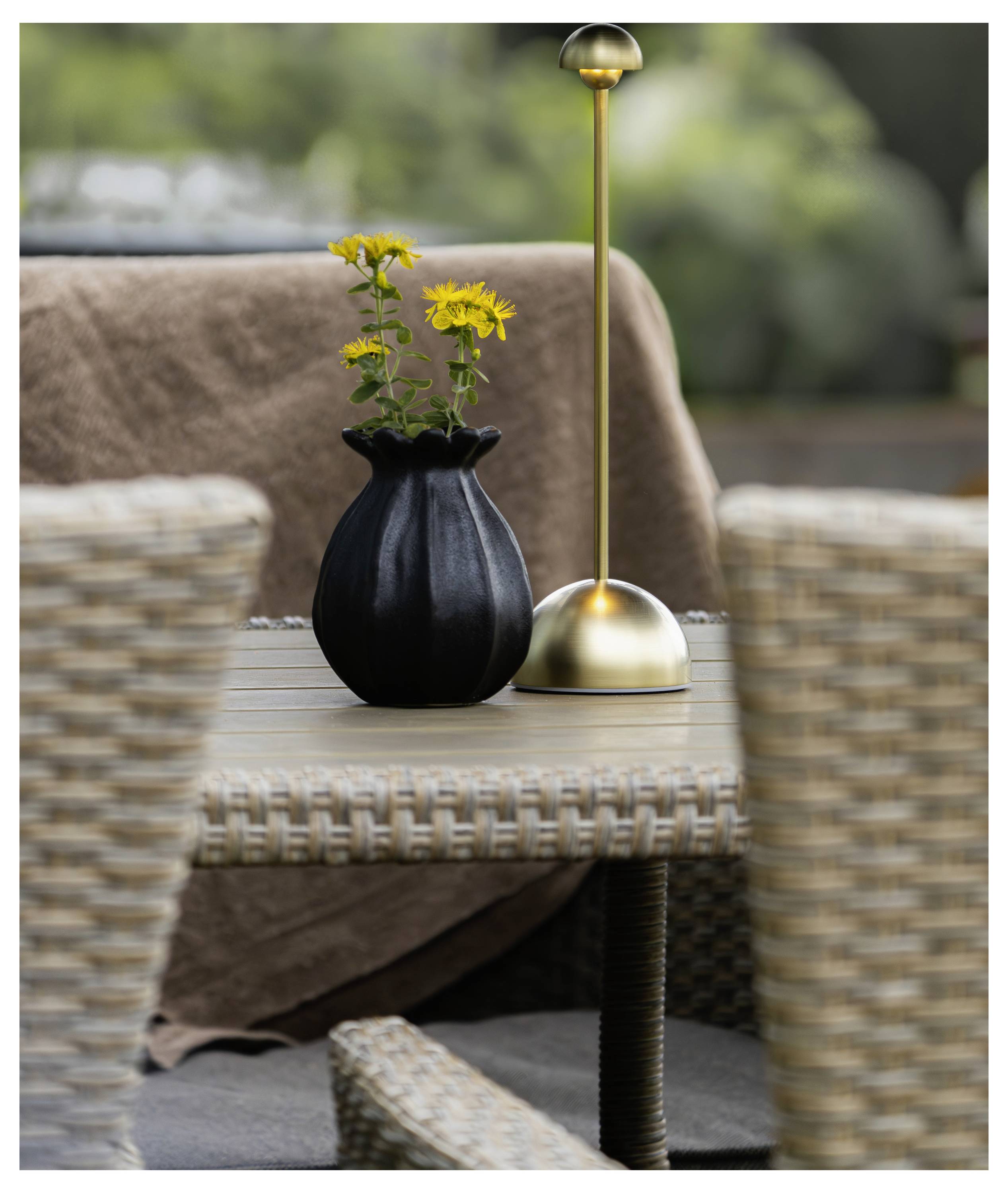 Vaso nero con fiori gialli e una lampada da tavolo in ottone su un tavolo intrecciato da esterno, con sedie in vimini e un plaid marrone sullo sfondo.