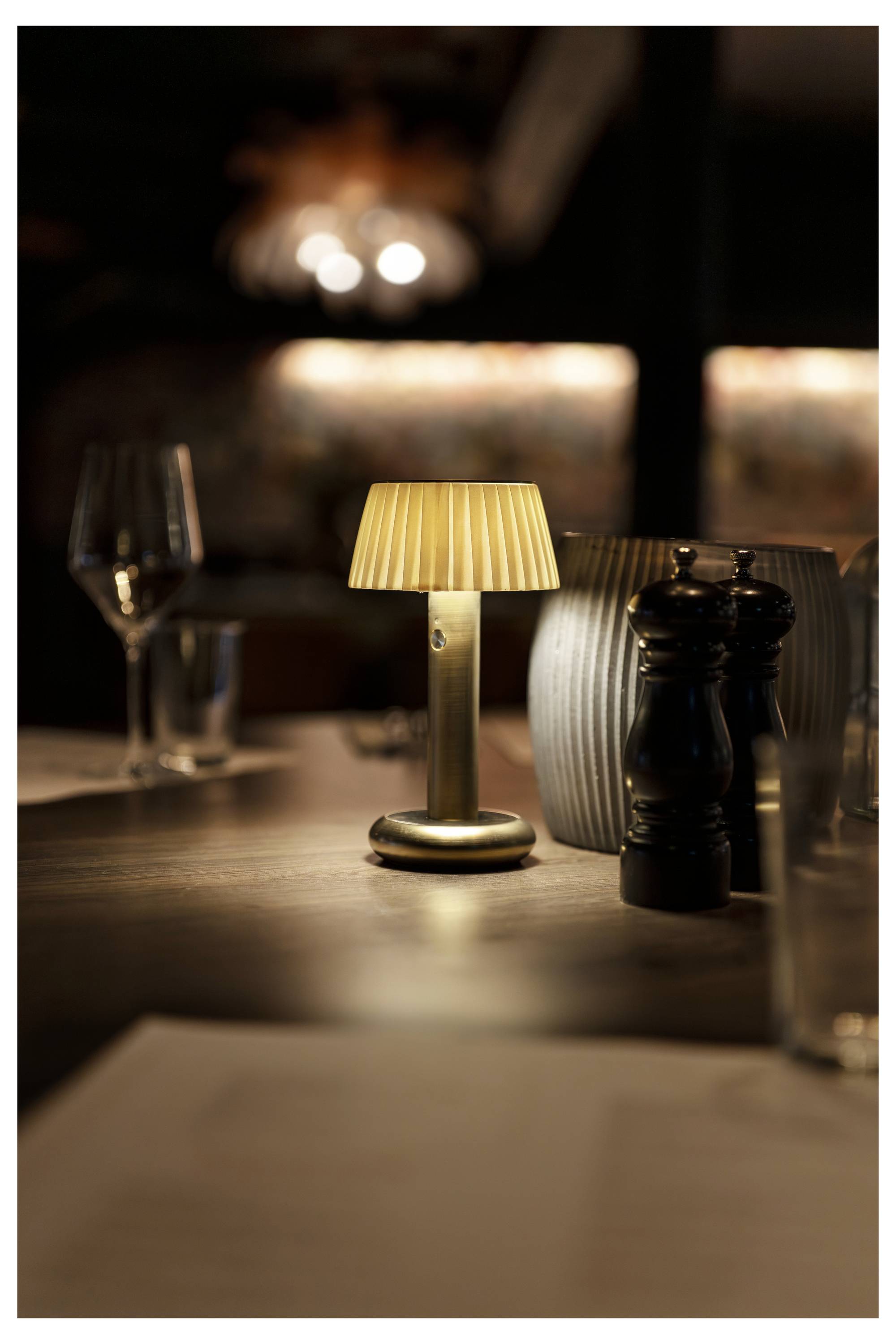 Un tavolo di ristorante illuminato da una luce calda con una piccola lampada, bicchieri di vino e saliera e pepiera sfocati dolcemente.
