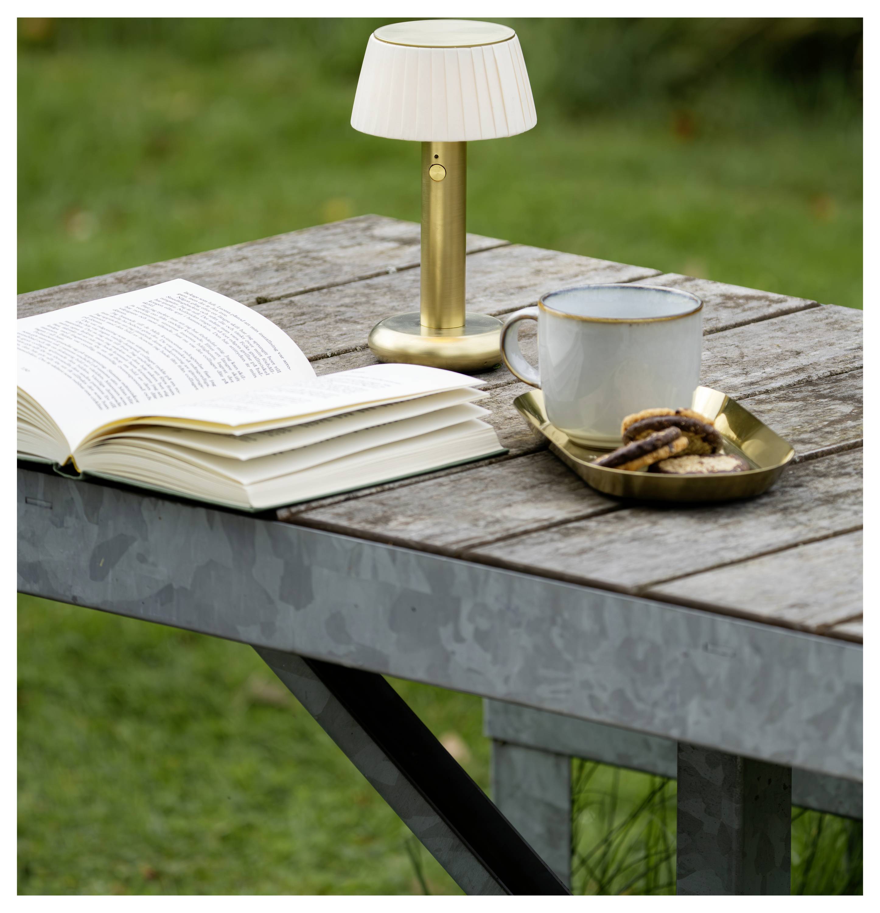 Tavolo da picnic all'aperto con un libro aperto, una piccola lampada, una tazza di tè e un piatto di biscotti sul piano del tavolo.