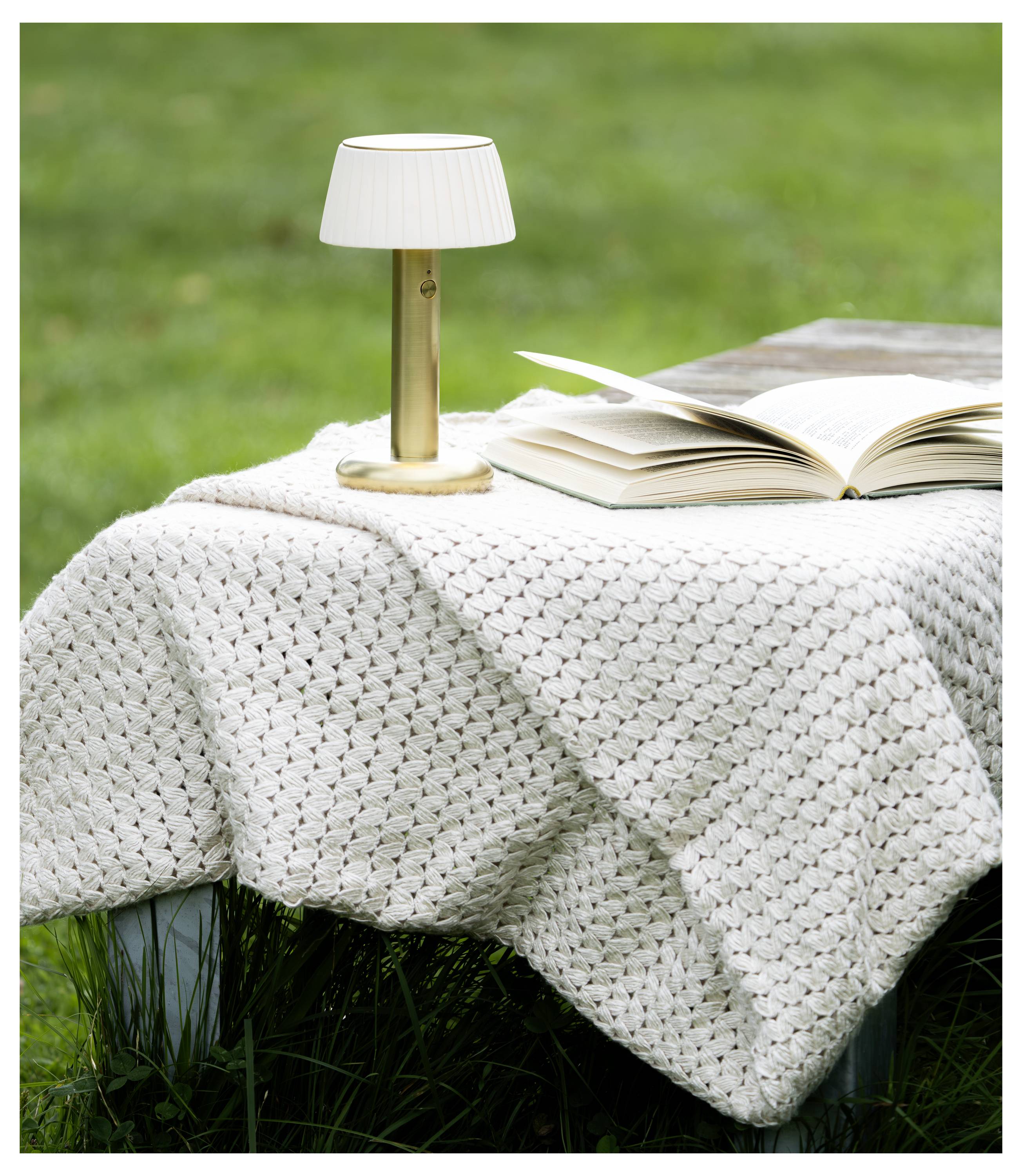 Libro aperto e piccola lampada da tavolo su una coperta in lana bianca lavorata a maglia su un tavolo all'aperto sull'erba.