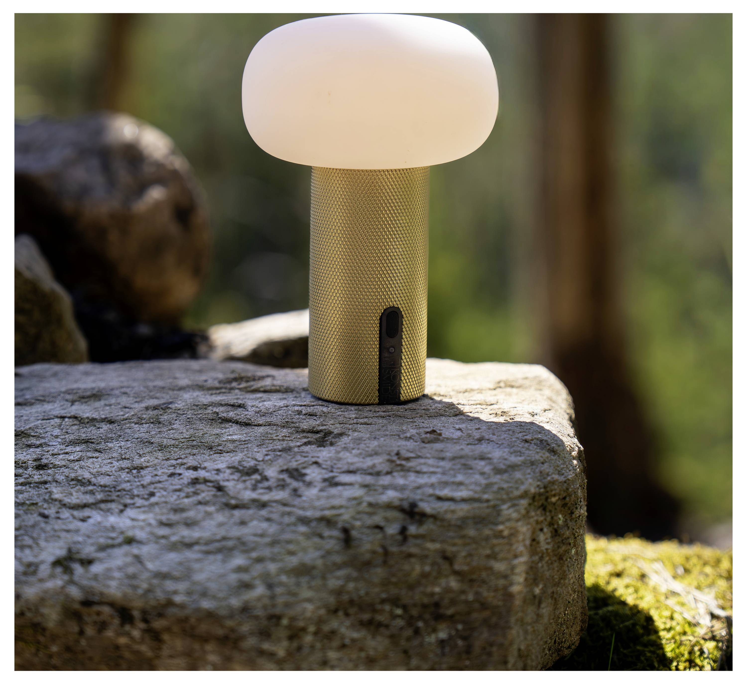 Piccola lampada portatile con paralume bianco luminoso e base cilindrica dorata che riposa su una pietra all'aperto.
