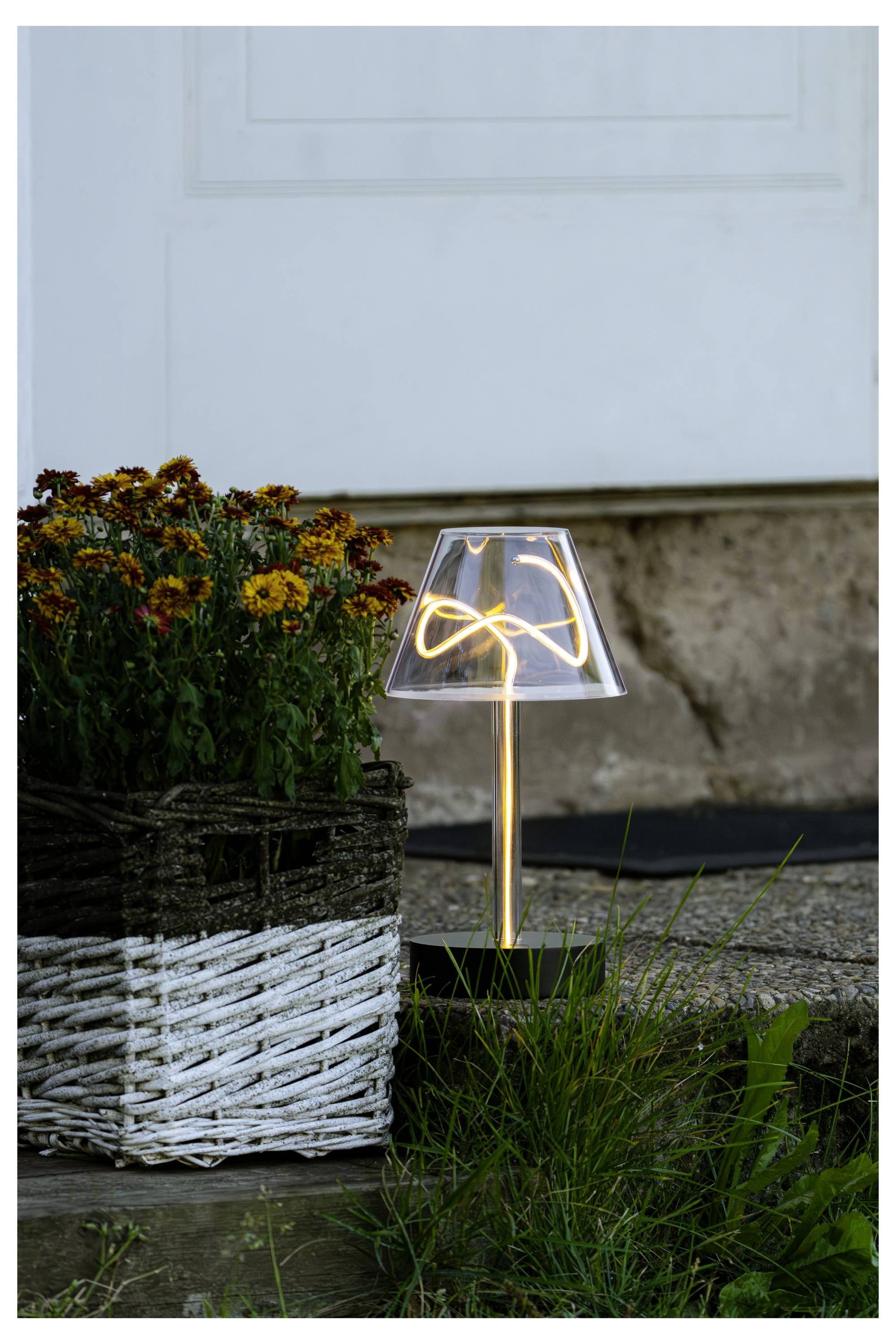 Una piccola lampada da tavolo luminosa accanto a un vaso in vimini bianco con fiori e erba, posizionata su una soglia davanti a una porta bianca.