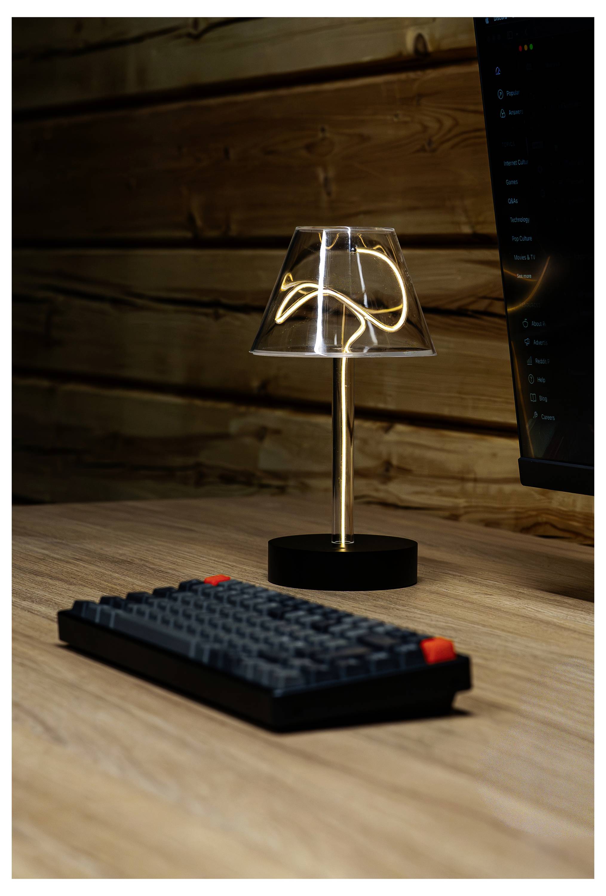 Una piccola lampada da scrivania moderna con paralume riflettente brilla su una scrivania in legno accanto a una tastiera compatta e a un monitor del computer.