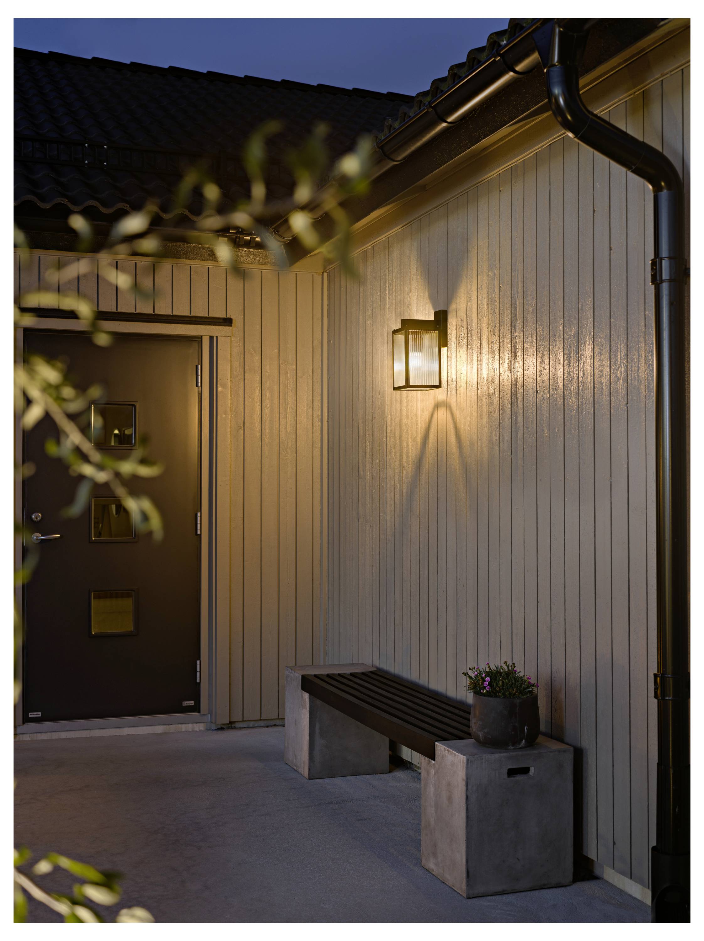 Ingresso esterno della casa al crepuscolo con una lanterna da parete illuminata, porta nera e panchina con pianta in vaso contro il rivestimento verticale.
