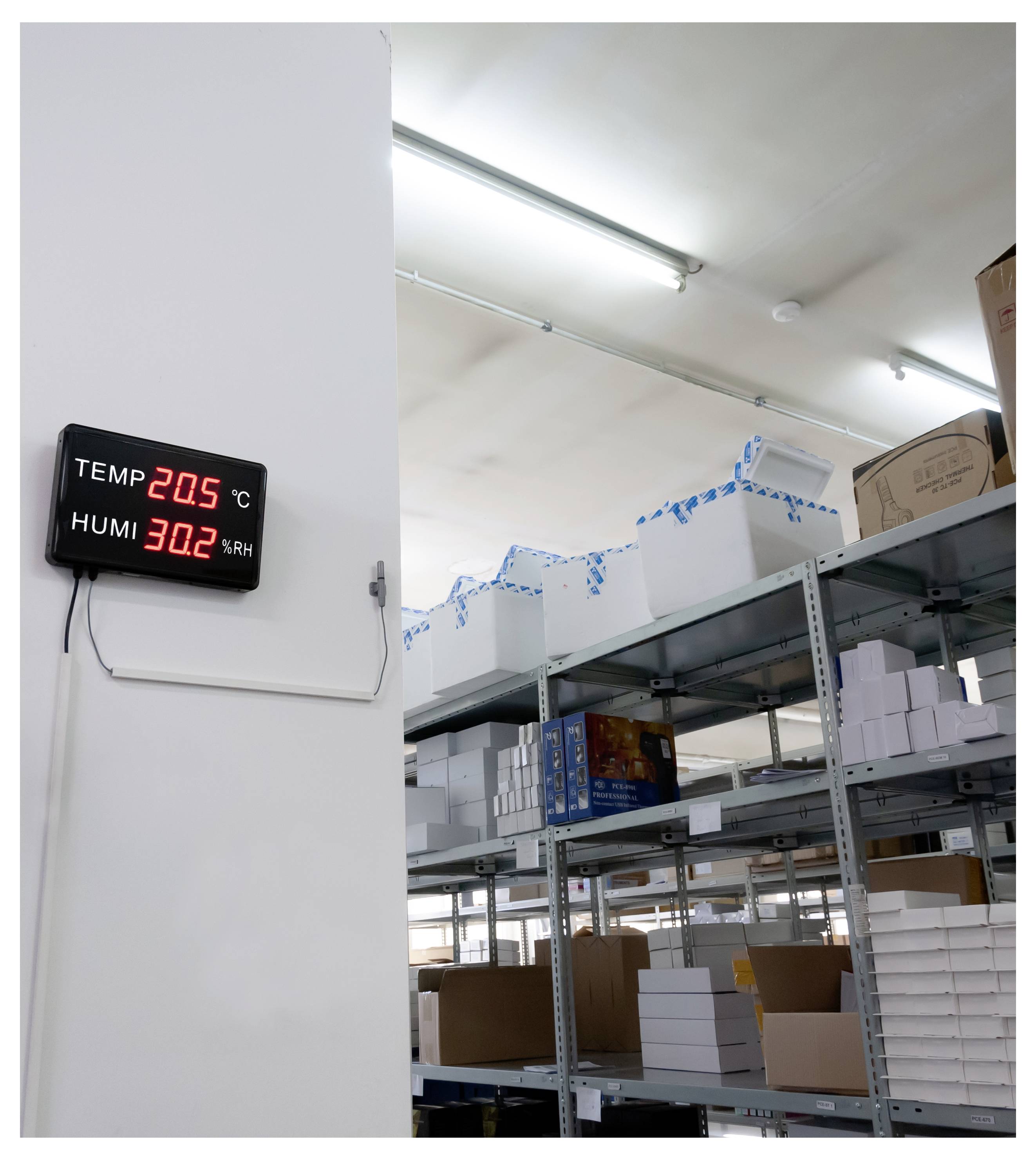 Magazzino con sala di stoccaggio e parete con display che mostra una temperatura di 20,5°C e un'umidità relativa del 30,2%, accanto a scaffali con scatole imballate.