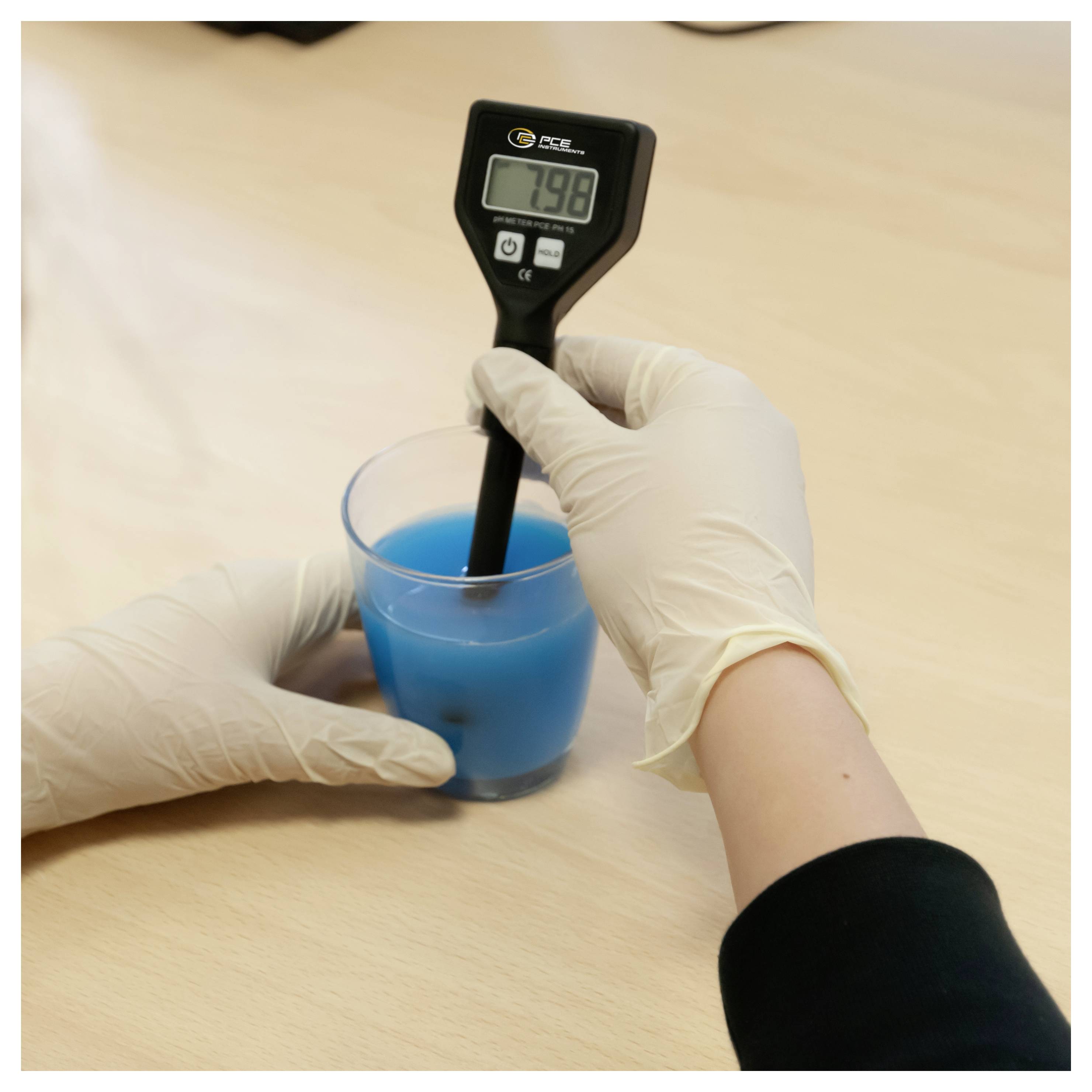 Le mani guantate utilizzano un misuratore digitale per misurare un liquido blu in una tazza trasparente su un tavolo.