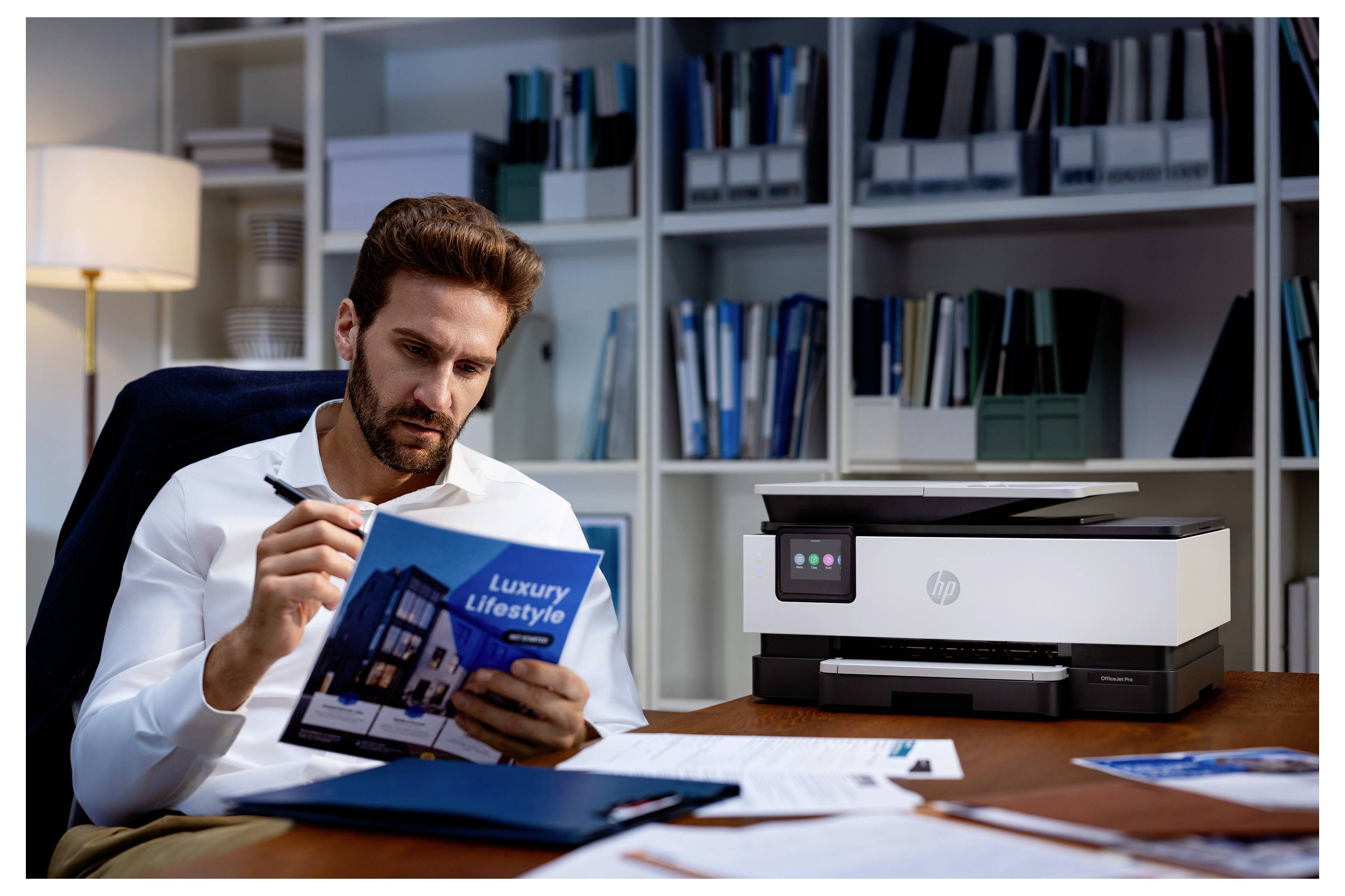Uomo in un ufficio studia una brochure "Stile di Vita Lussuoso" accanto a una stampante, con carte e cartelle sulla scrivania.