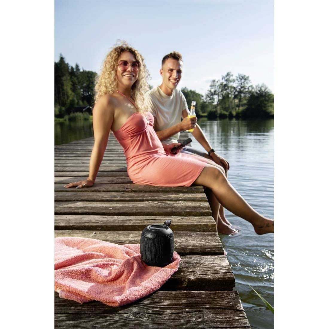 Due persone si rilassano su un molo di legno vicino a un lago; una donna in un abito rosa è seduta davanti, mentre un uomo dietro di lei tiene in mano una bevanda.