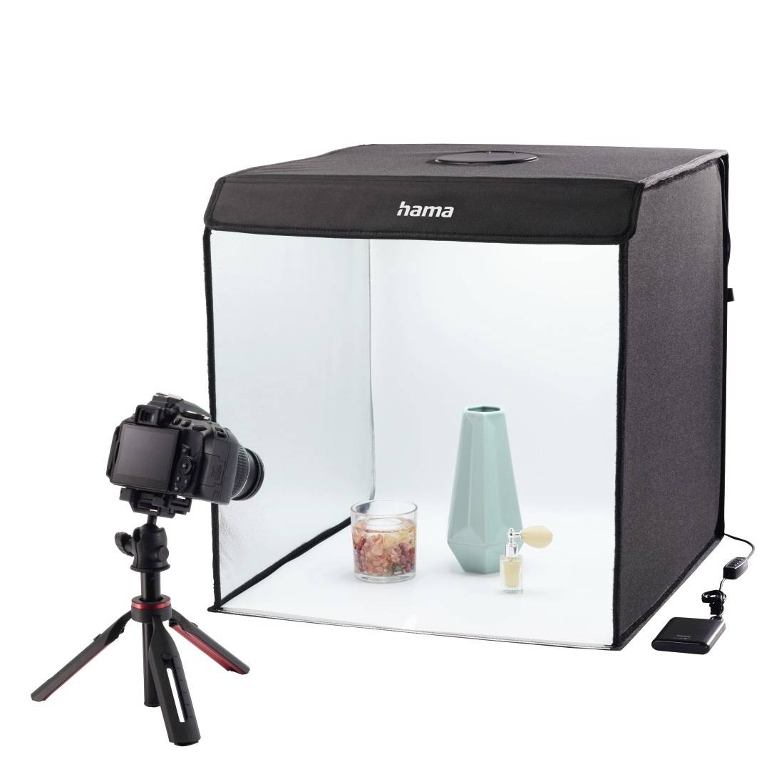 Fotocamera su treppiede che fotografa un prodotto all'interno di un piccolo studio lightbox bianco con un'etichetta 'hama', su uno sfondo bianco.