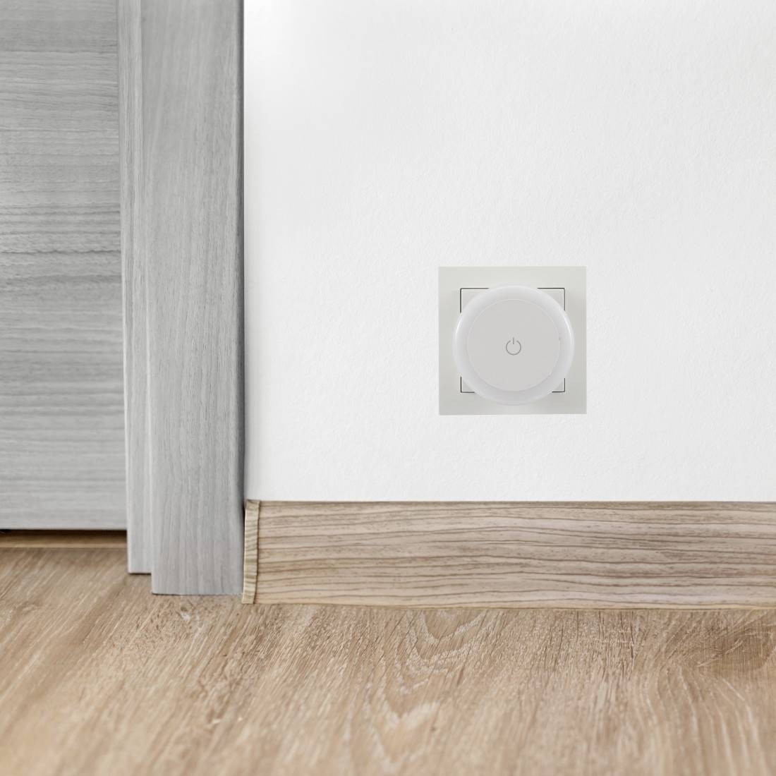 Parete bianca con un piccolo pannello di controllo/pulsante di alimentazione quadrato sopra un pavimento in legno e battiscopa vicino a una porta o bordo di un armadio.