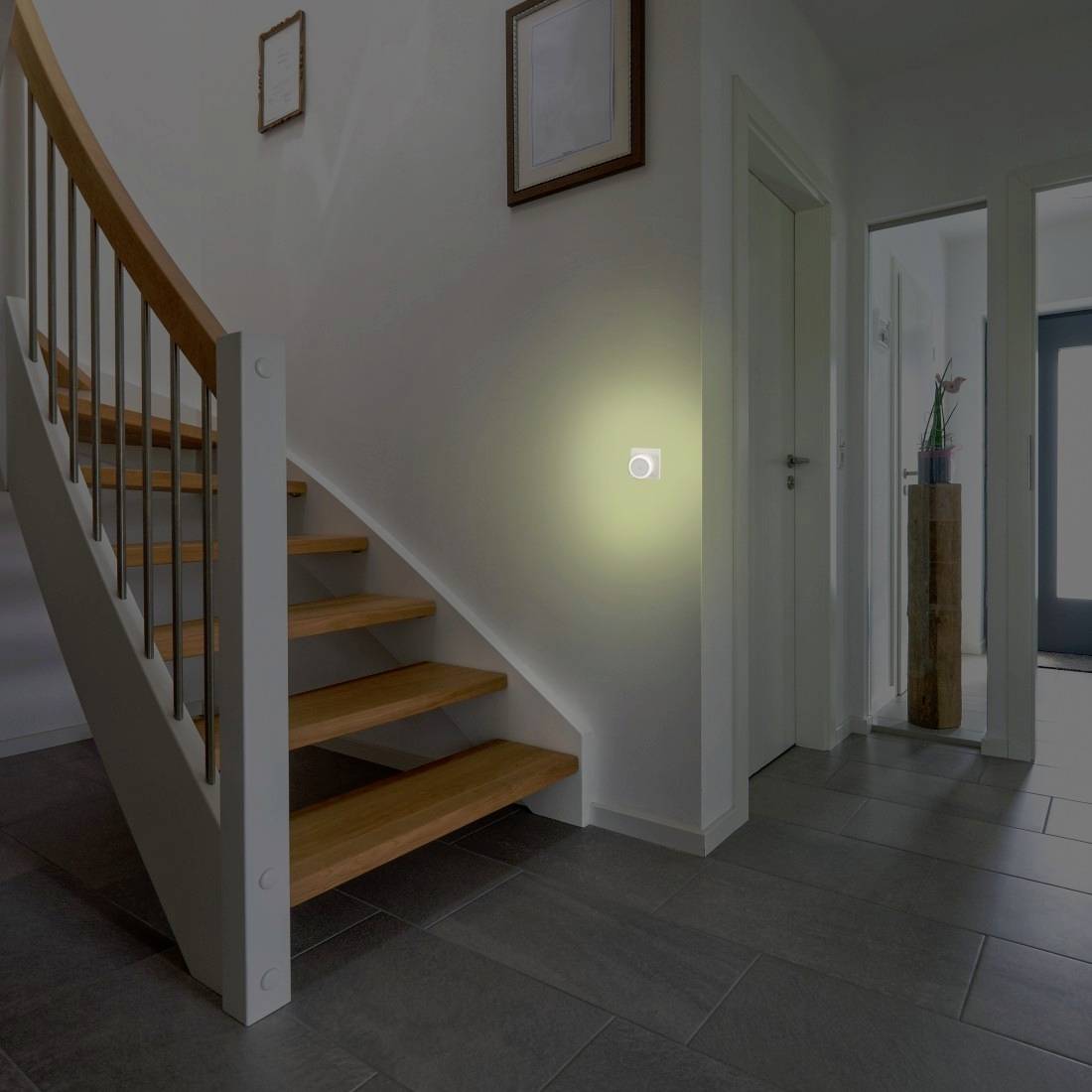Corridoio con scala in legno e una lampada da parete che brilla su un muro bianco, accanto alle porte che conducono ad altre stanze.
