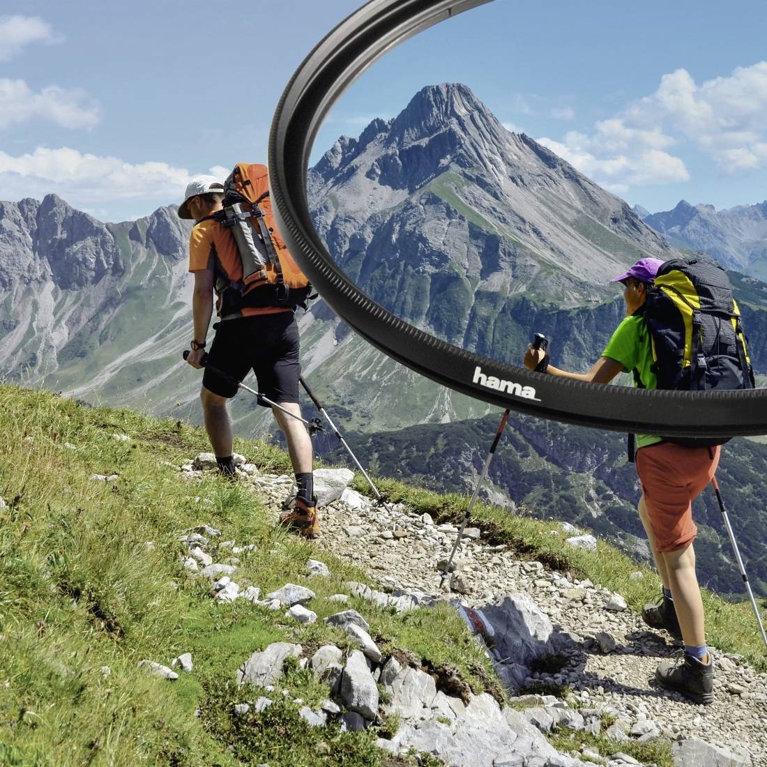 Due escursionisti con zaini e bastoni da trekking camminano lungo un sentiero di montagna, incorniciati da un grande filtro a lente circolare, con picchi drammatici sullo sfondo.