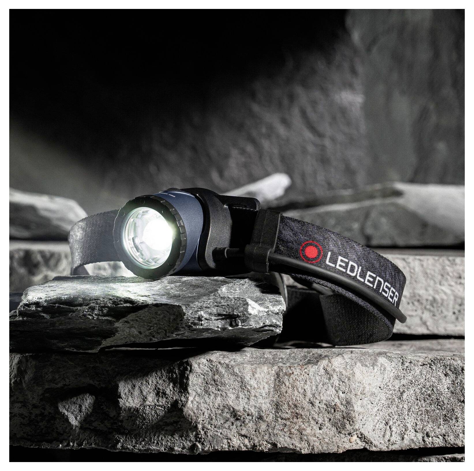 Lampada frontale LEDLENSER accesa appoggiata su rocce impilate, che mostra un fascio luminoso brillante e la cinghia regolabile in un ambiente esterno aspro.