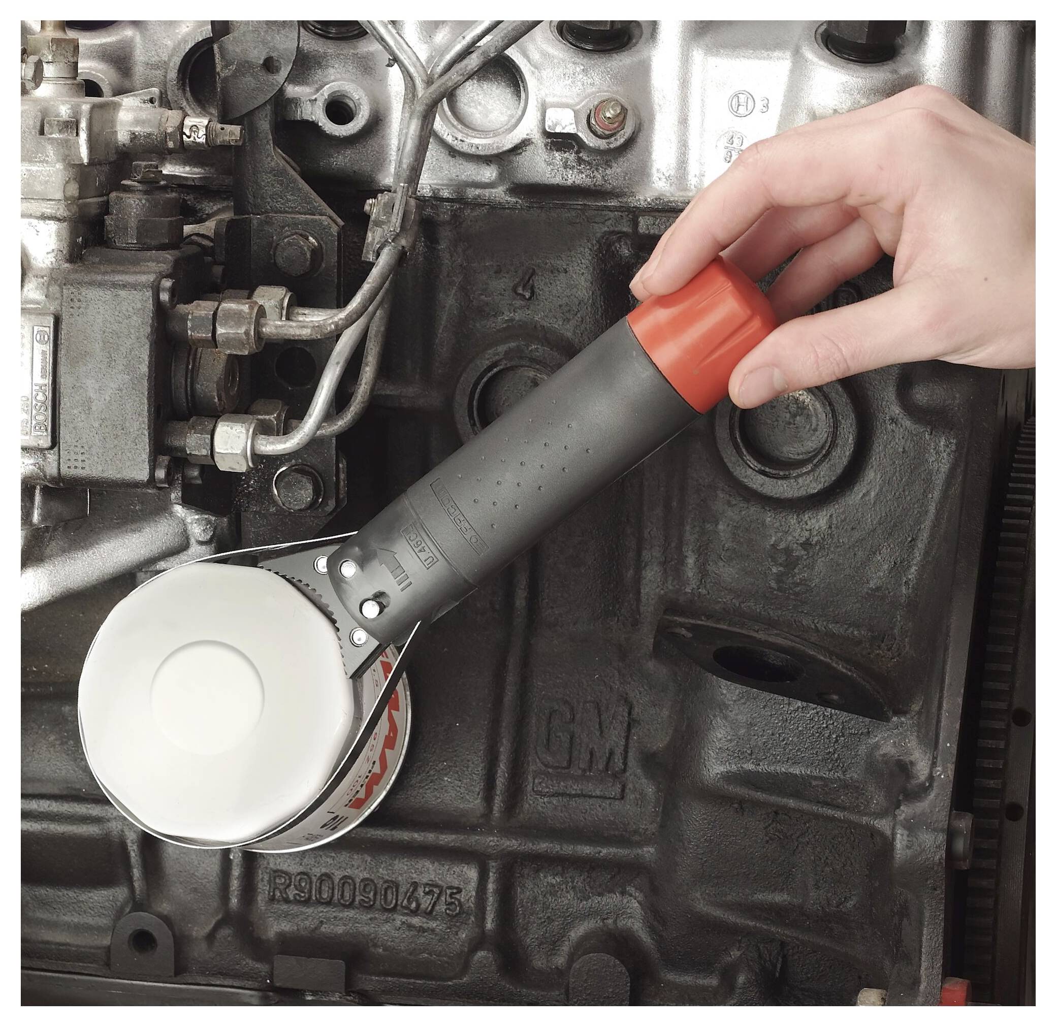 Una mano tiene una lampada a torcia con manico lungo all'interno di un vano motore buio, illuminando il blocco motore e i componenti metallici circostanti.