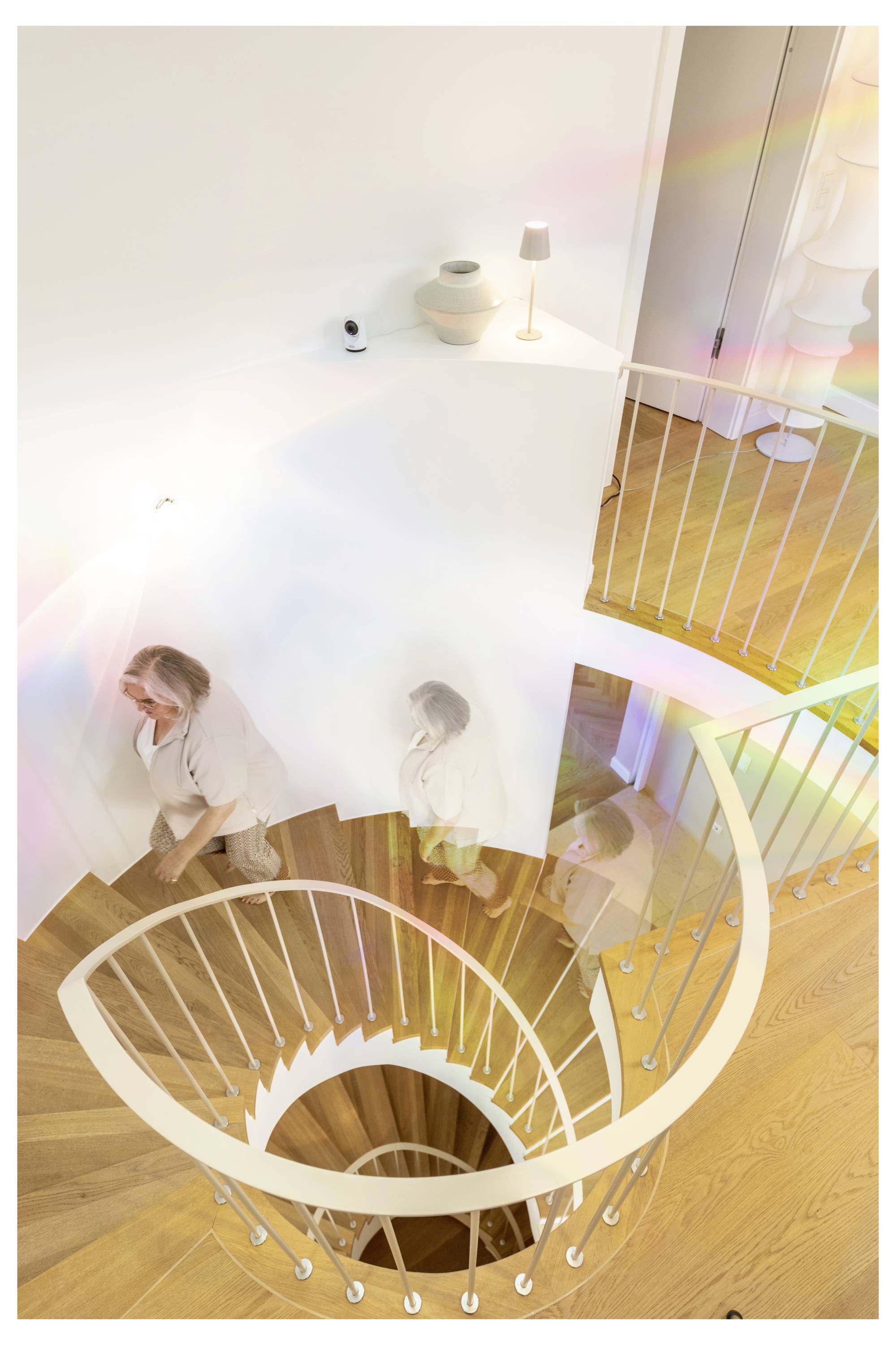 Vista dall'alto di una scala bianca curva con una persona che scende; pavimenti in legno caldo e luminoso arredamento interno visibile.