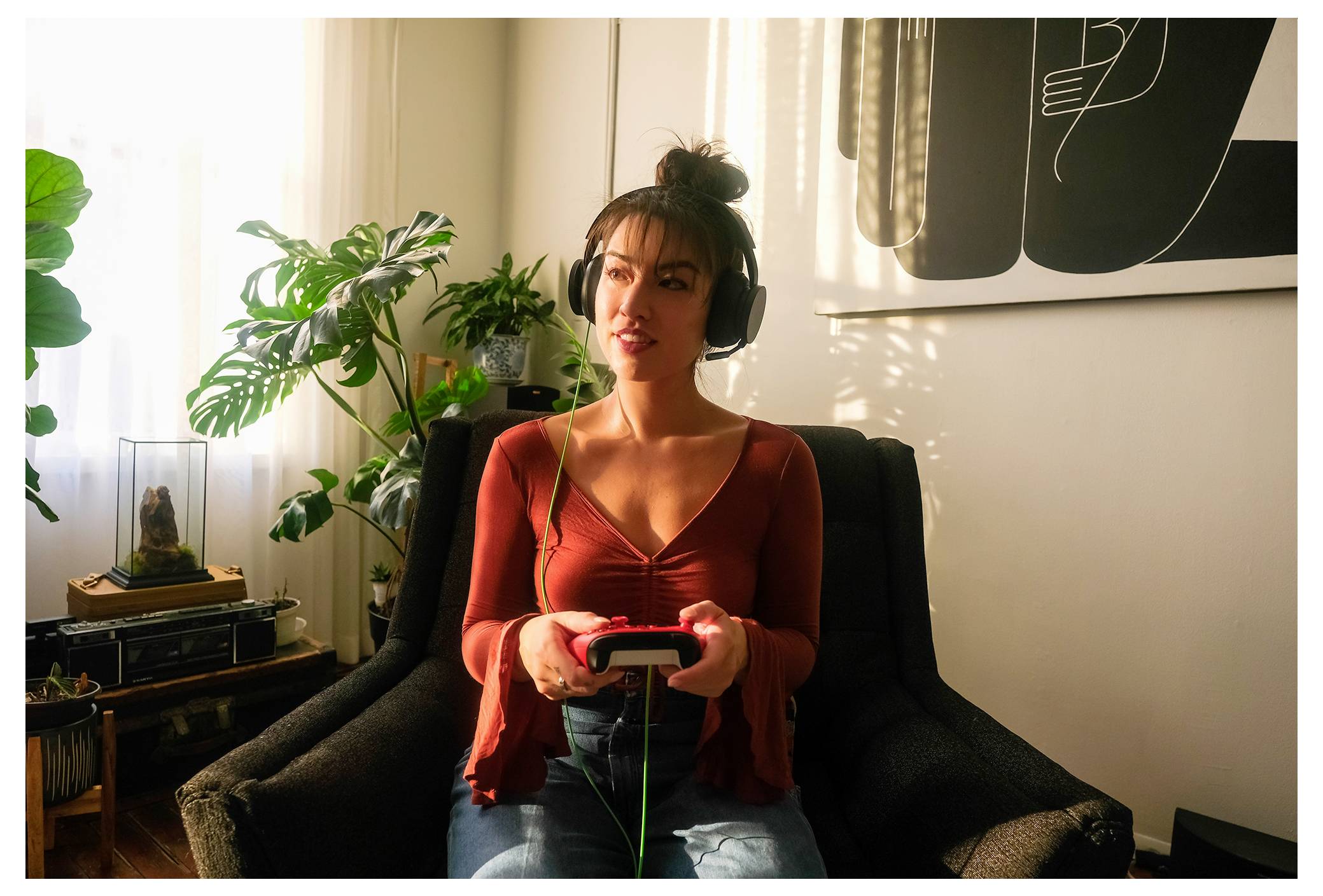Una donna che indossa cuffie gioca a un videogioco mentre è seduta su una sedia in un soggiorno illuminato dal sole.