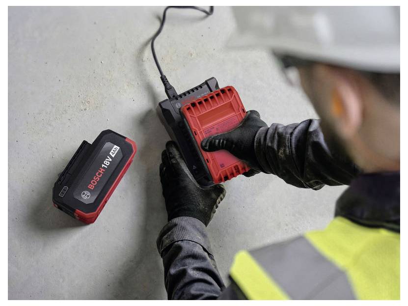 Un operaio in equipaggiamento di sicurezza collega un caricabatterie Bosch rosso a 18V a una presa a muro accanto a un pacco batteria.